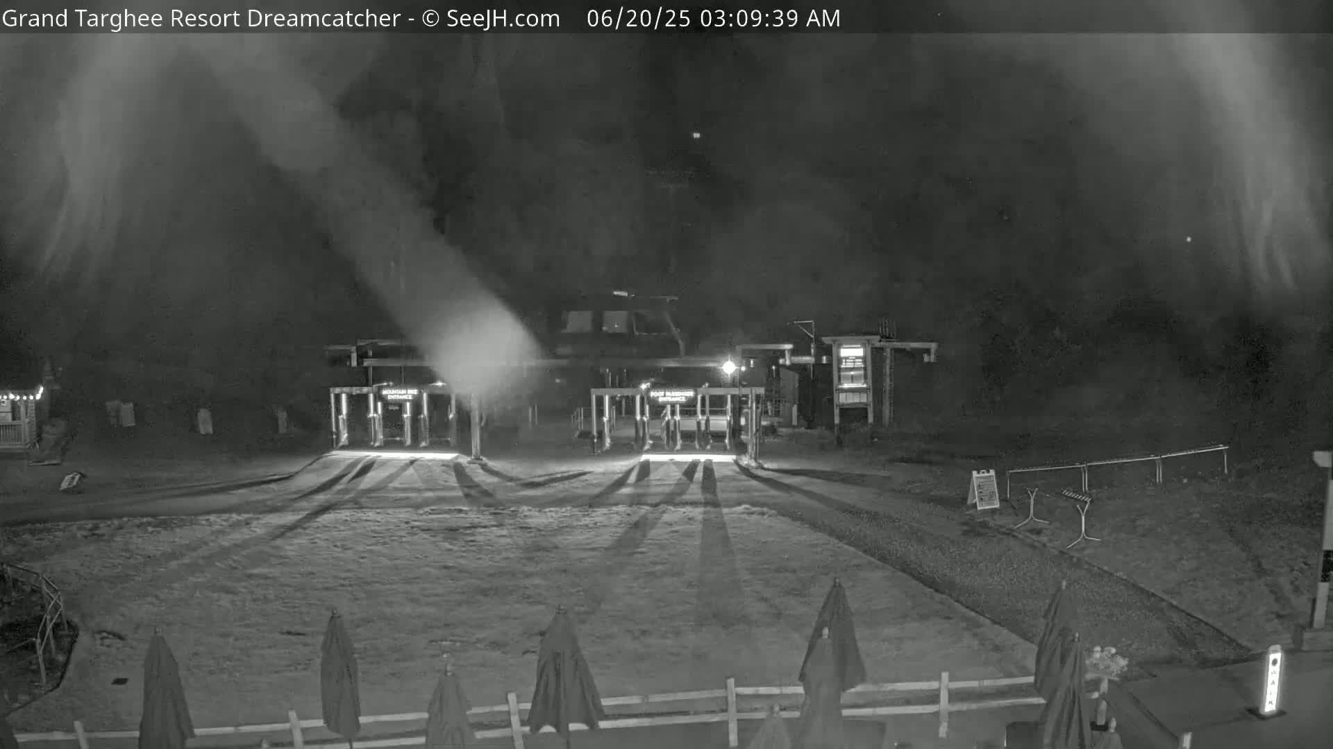 A nighttime, snow-covered area with a building and entry gates, illuminated by lights, with plumes of snow or mist emanating from the building.