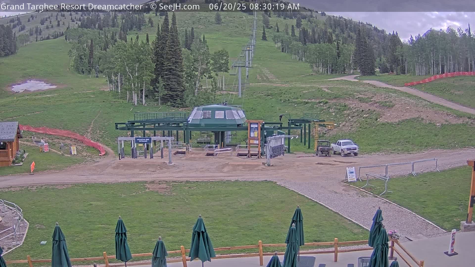 A mountainside ski lift base area with overcast skies, a mostly green grassy hillside, and several dark-green patio umbrellas.