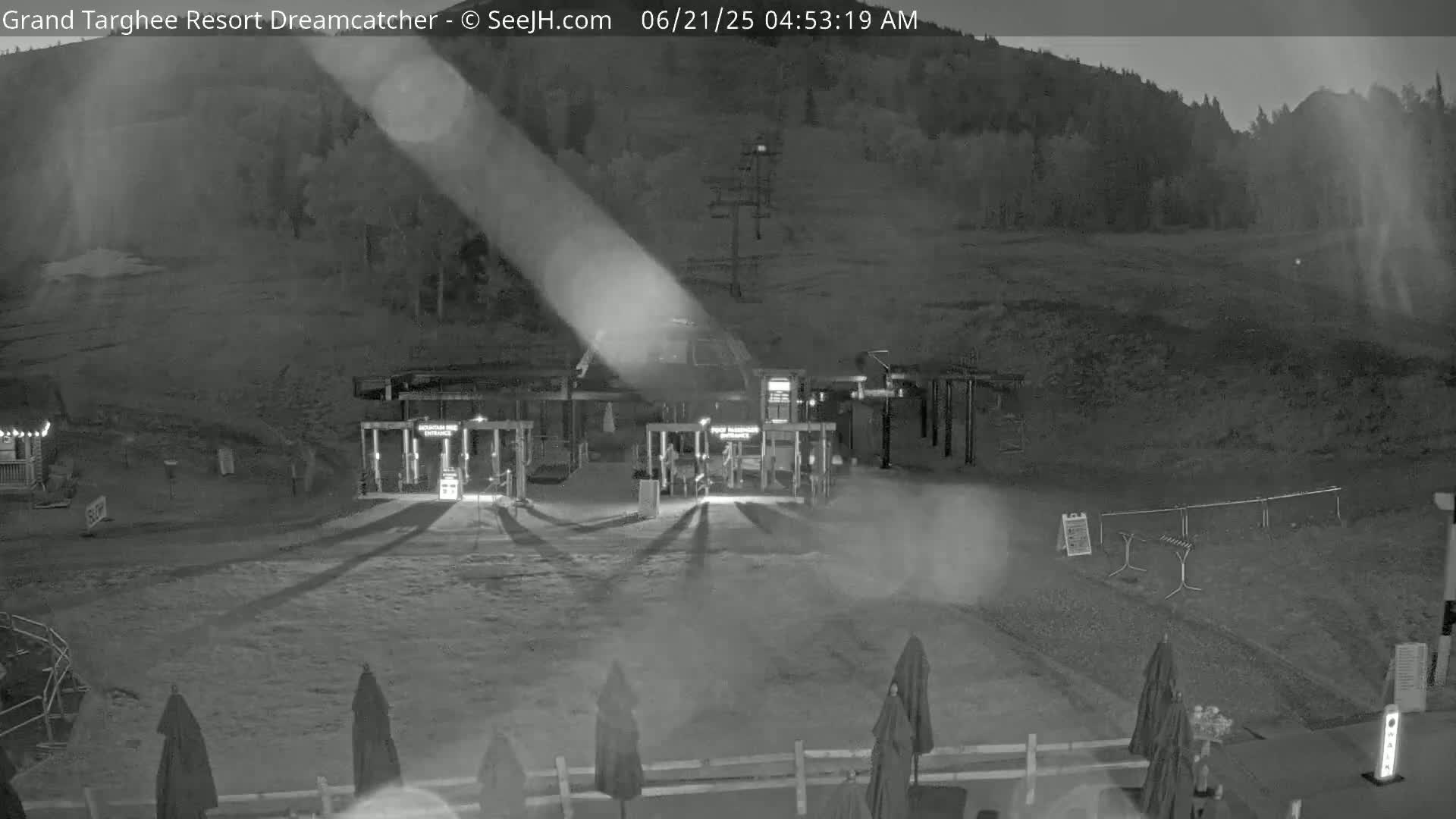 A nighttime, low-light view of a ski lift base area with several closed gates, some outdoor furniture, and a hill covered in trees in the background.