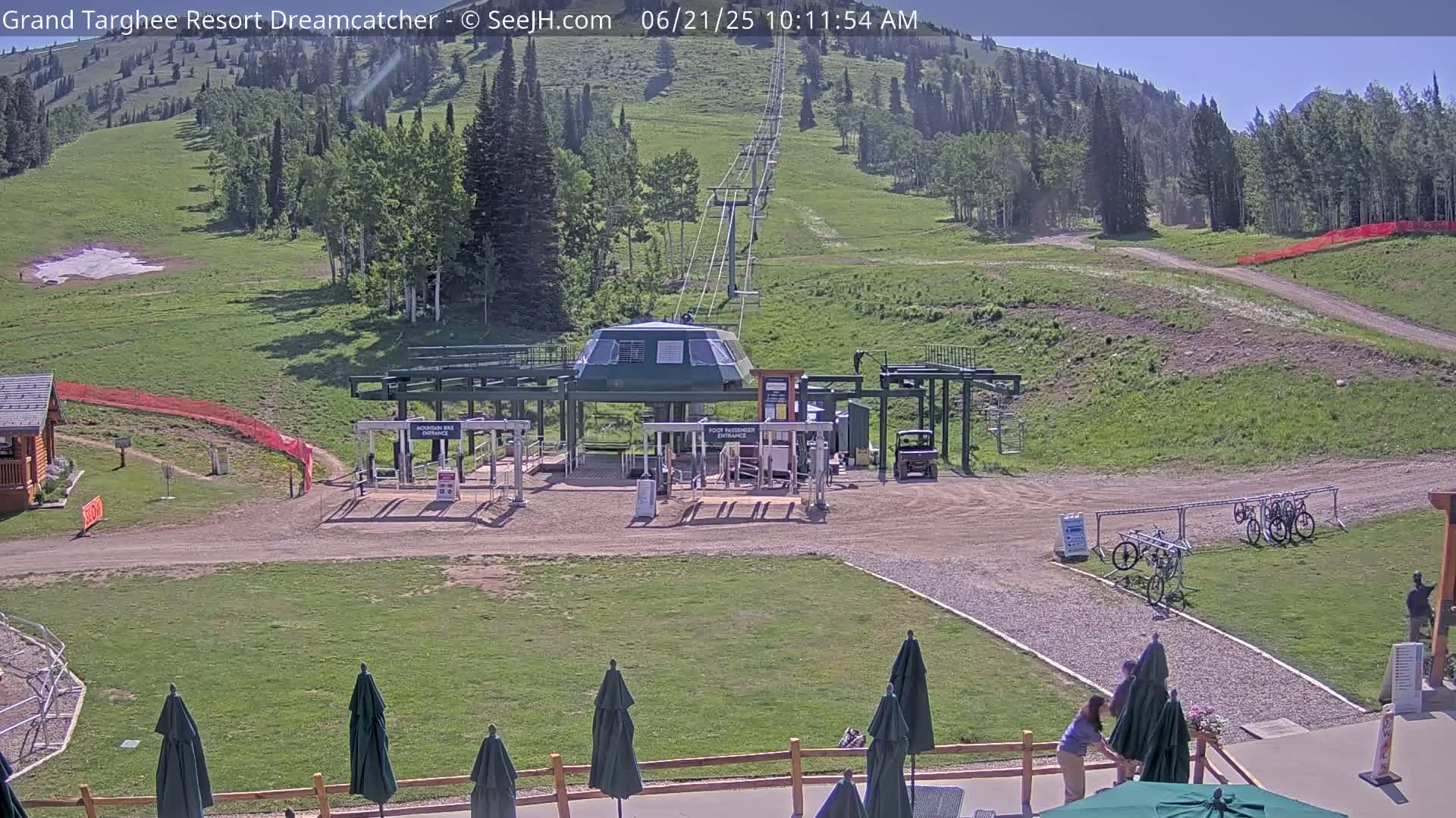 A mountainside chairlift base station with several parked bicycles and people preparing patio umbrellas under a sunny sky.