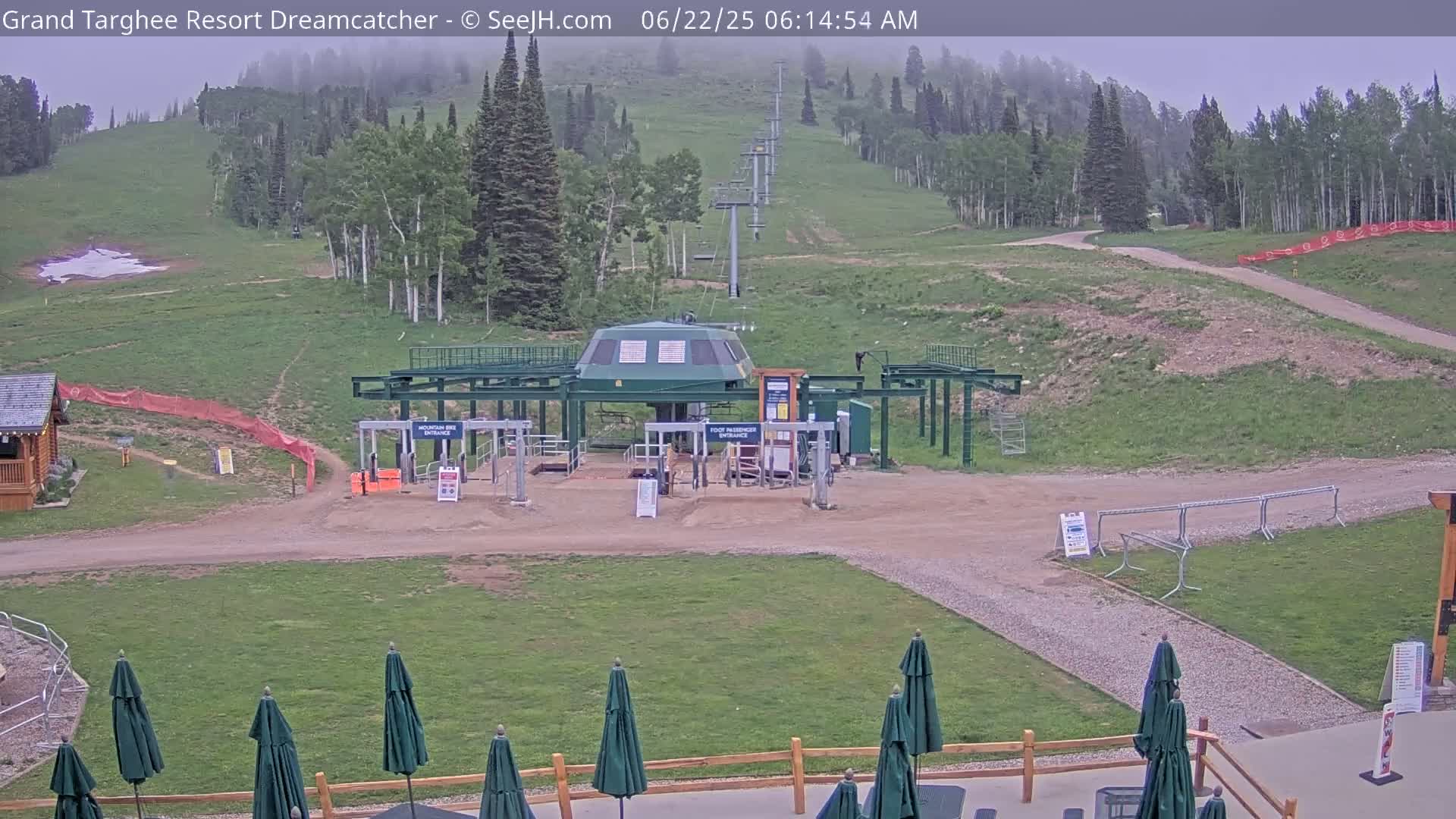A ski lift base station sits on a grassy hill under an overcast sky, with several closed umbrellas and a wooden fence visible in the foreground.