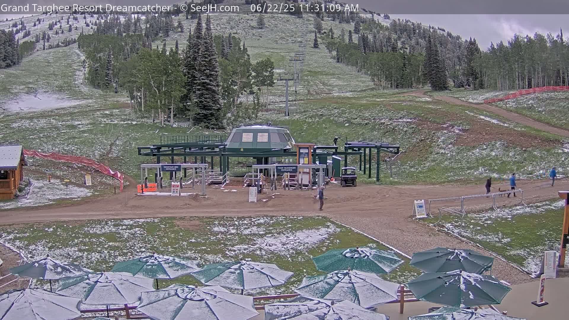 A ski lift base area is lightly dusted with snow, with several people walking nearby and closed green umbrellas covered in snow in the foreground.