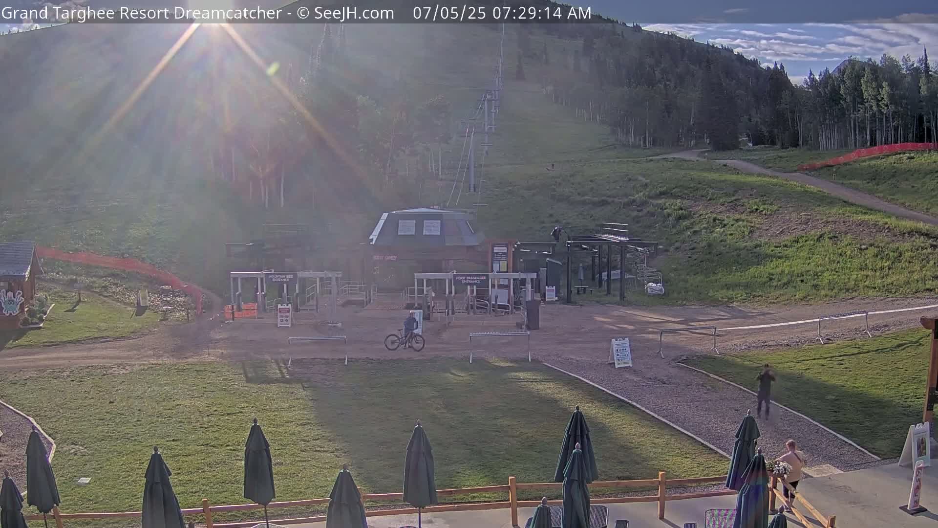 A sunny morning scene at a mountain resort shows a chairlift, a base area with a few people, and a mountainside with grassy slopes.