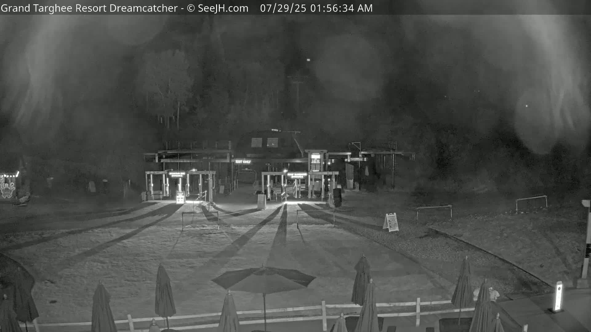 A nighttime, foggy view of a gated entrance to a building, with several closed umbrellas and long shadows cast across a paved area.