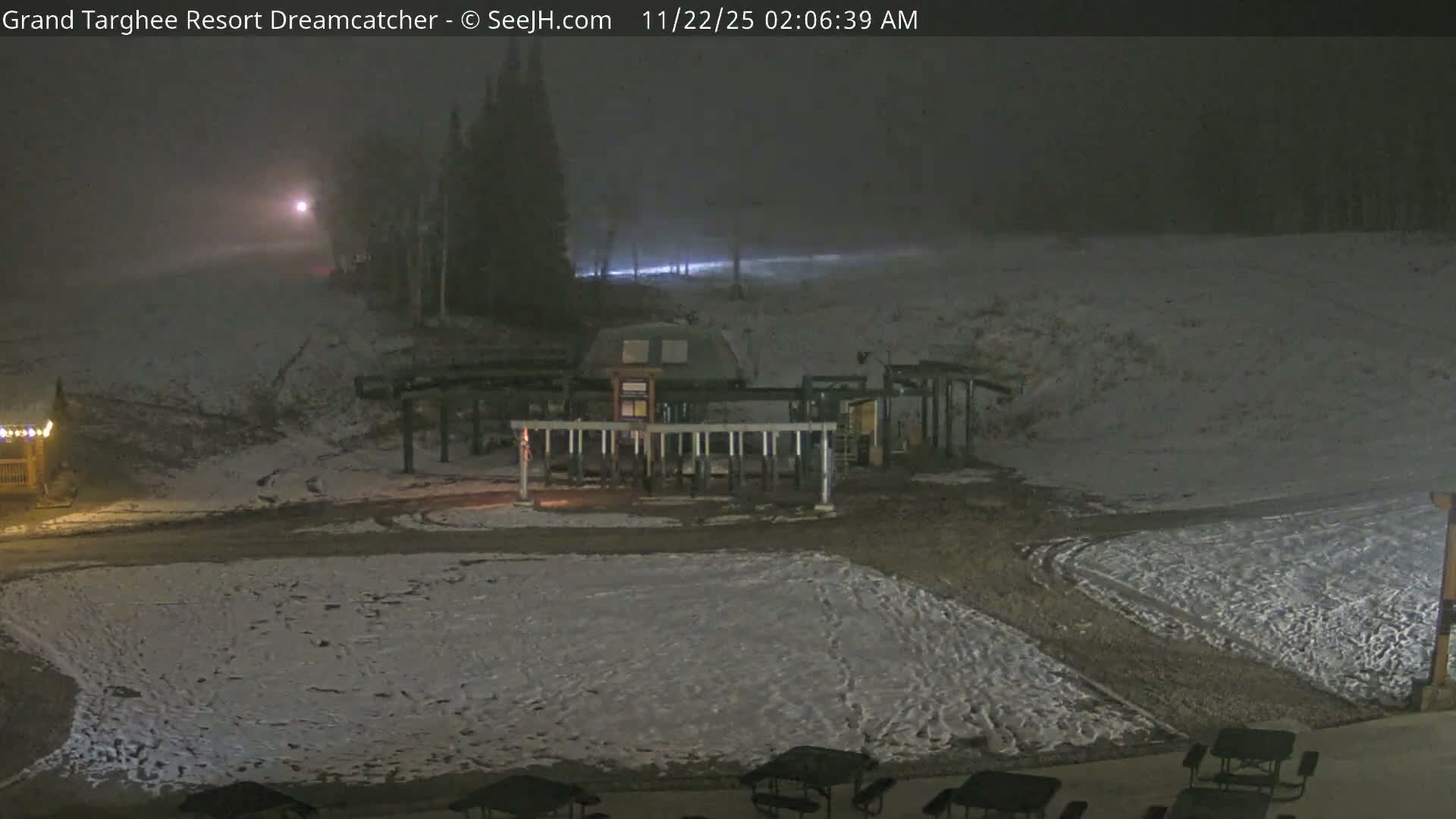 A snow-dusted ski resort is visible at night, featuring a lit ski lift station and scattered trees under foggy or overcast skies.