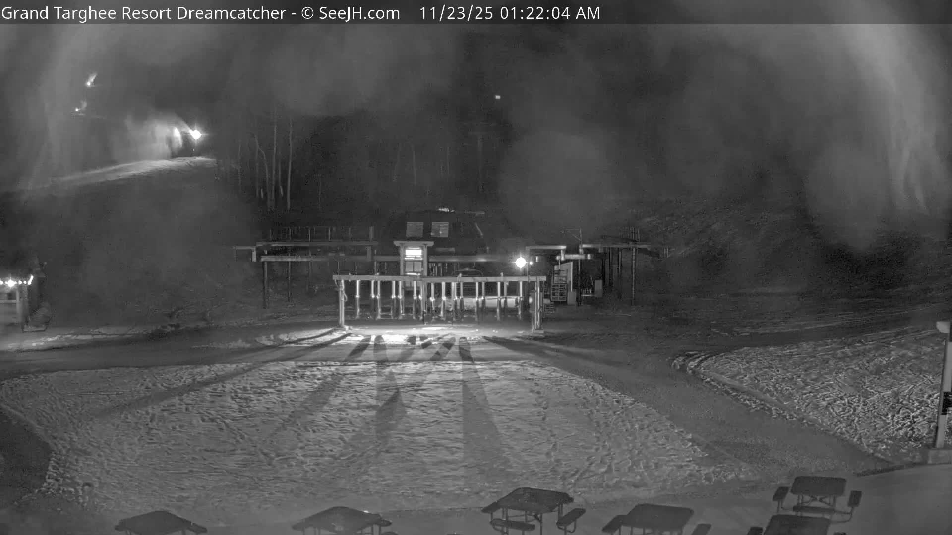 A cold, dark night at a ski resort features a brightly lit ski lift entrance and snow-covered ground in the foreground, with multiple snow-making machines actively creating snow on the slopes in the background.
