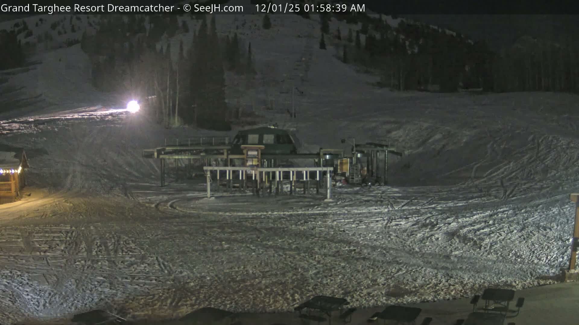 A nighttime scene at a snow-covered ski resort reveals a prominent chairlift station, well-used slopes crisscrossed with ski tracks, and artificial lights illuminating parts of the mountain under clear and cold winter conditions.