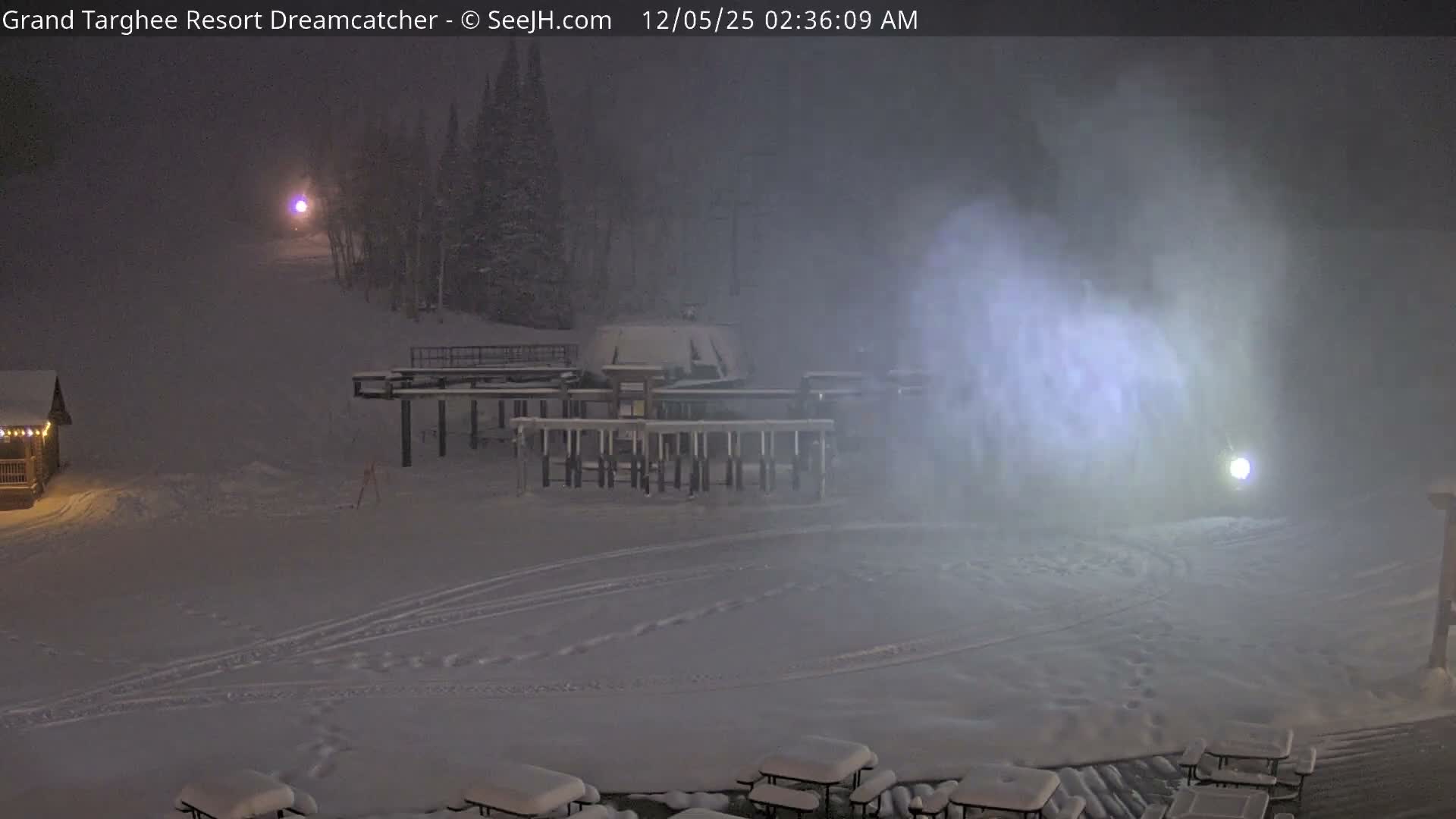 A snow-covered ski resort is seen at night, with fresh powder on the slopes and buildings, under a hazy atmosphere with visible falling snow illuminated by artificial lights.