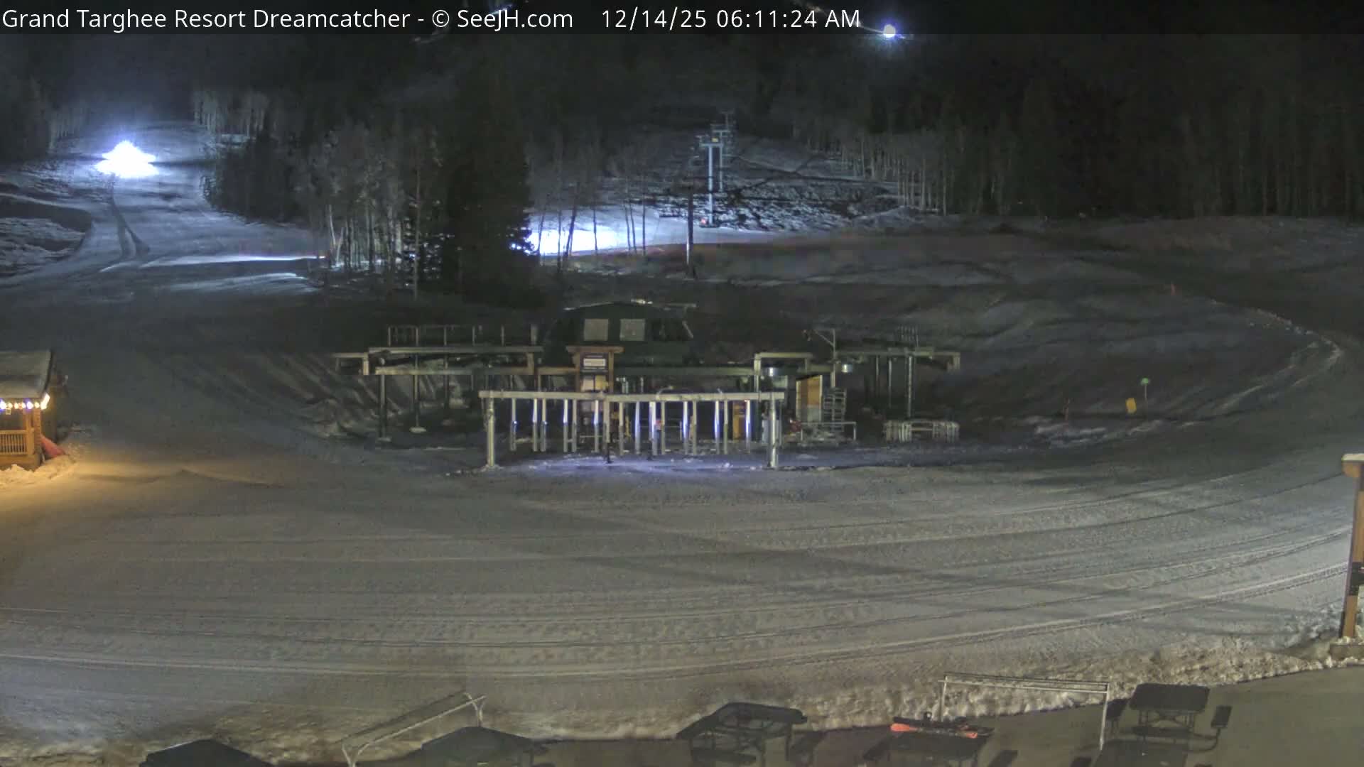 A snow-covered ski resort is seen at night, with fresh powder on the slopes and buildings, under a hazy atmosphere with visible falling snow illuminated by artificial lights.