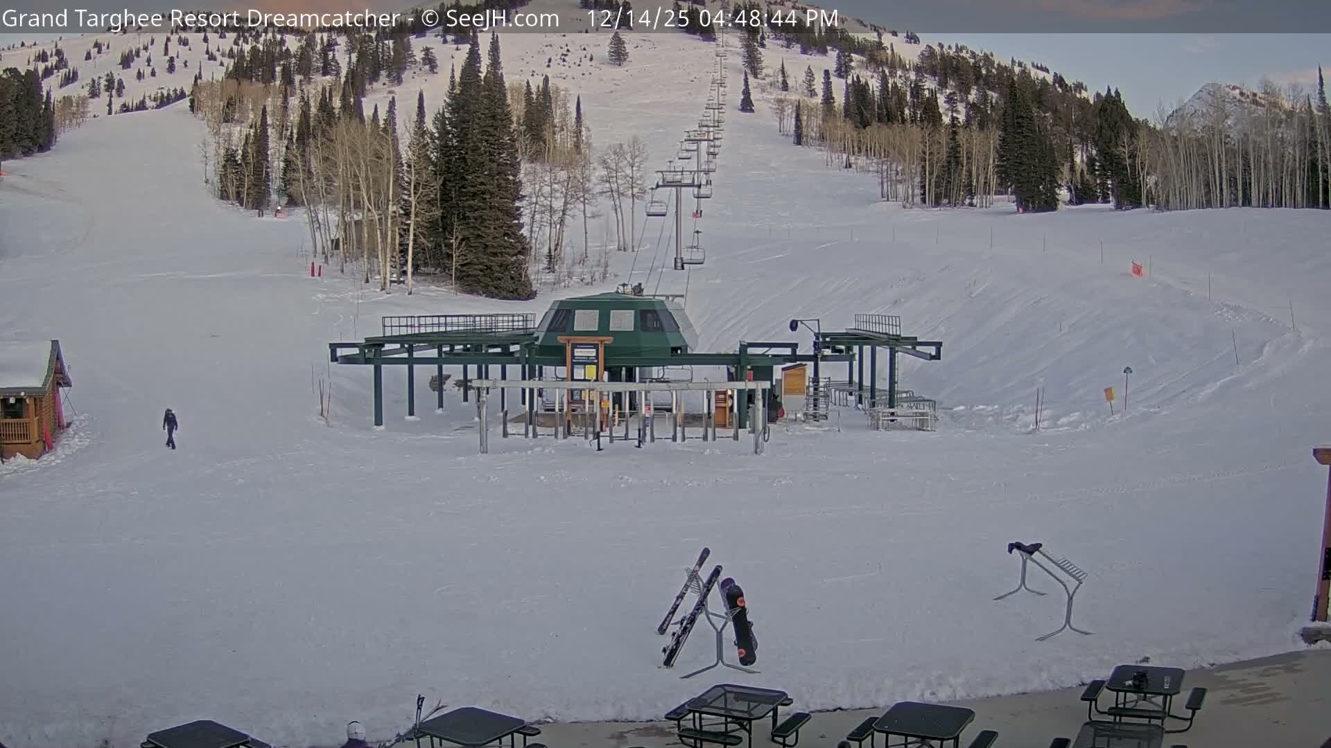 A snow-covered ski resort is seen at night, with fresh powder on the slopes and buildings, under a hazy atmosphere with visible falling snow illuminated by artificial lights.