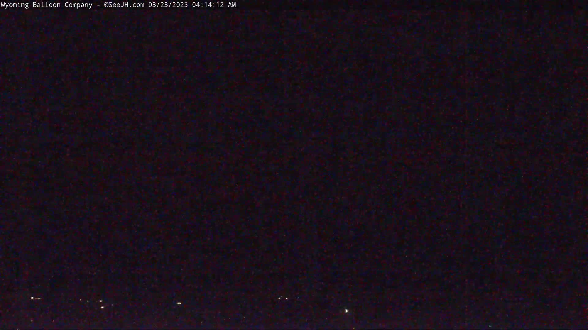 A dark, nighttime scene shows a few faint lights on the horizon under a mostly clear, dark sky.