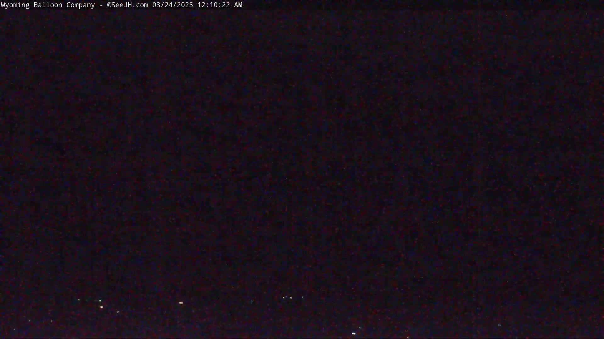 A nighttime, dark image shows a few faint lights on the horizon under a mostly clear, dark sky.