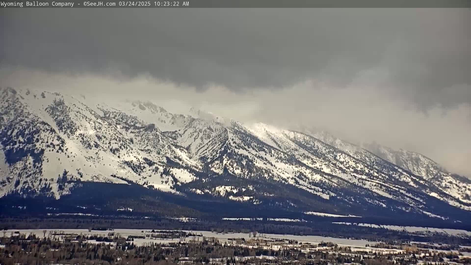 Snow-covered mountains under a cloudy, overcast sky overlook a valley with a town below.