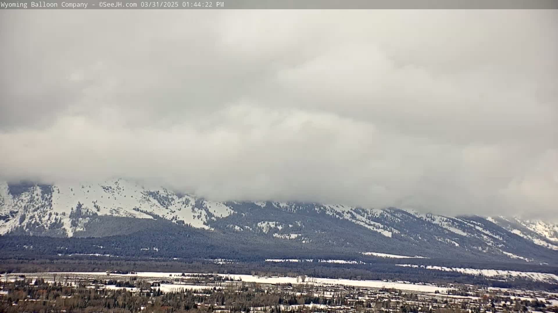 A snow-covered mountain range is visible under a cloudy sky, with a town or village situated in the valley below.