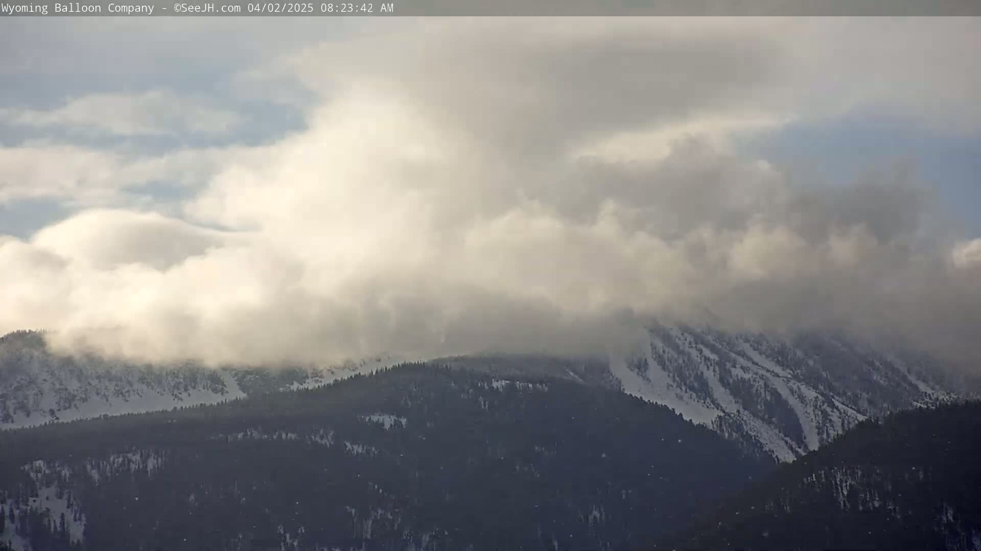 A snow-covered mountain range is partially obscured by a low layer of clouds under an overcast sky.