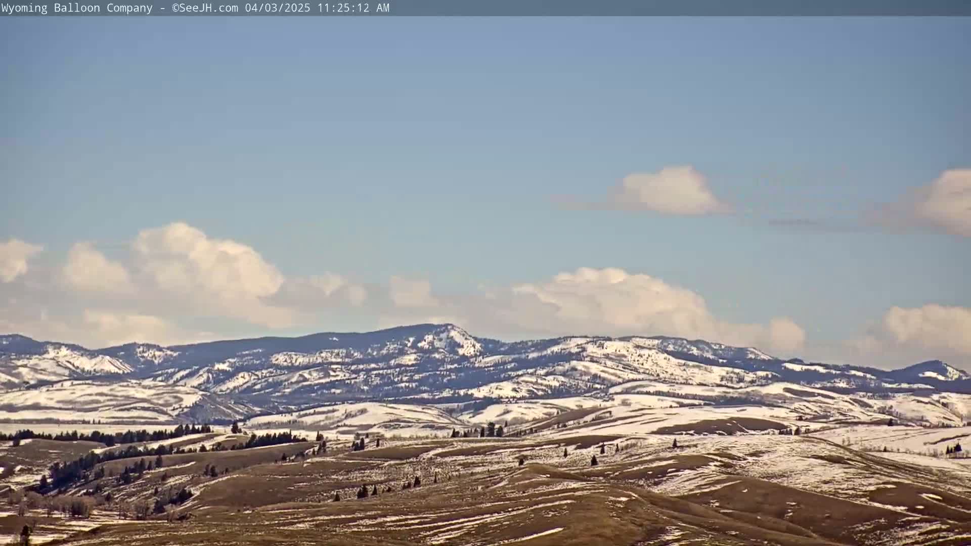 A mostly clear, slightly cloudy sky overlooks snow-dusted rolling hills and mountains.