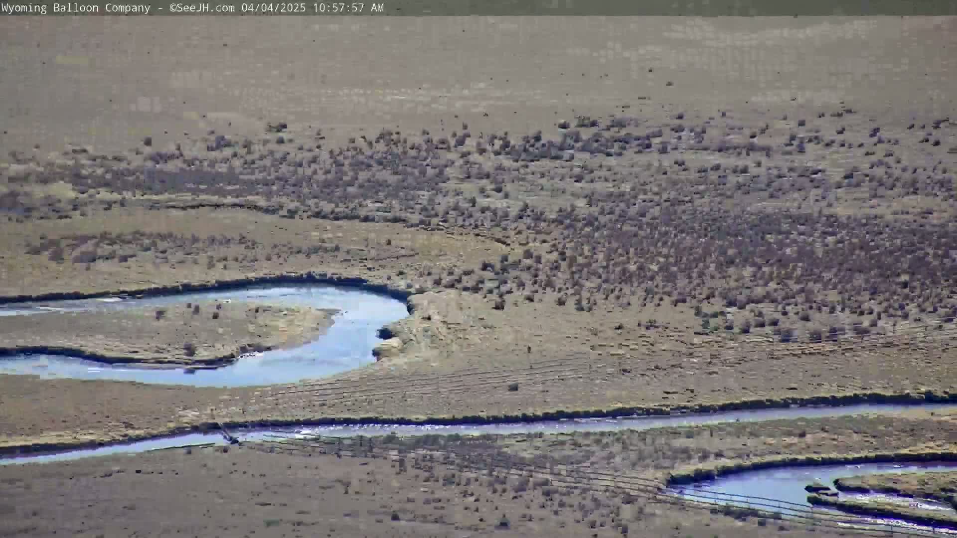 A winding river flows through a dry, sparsely vegetated landscape under sunny conditions.