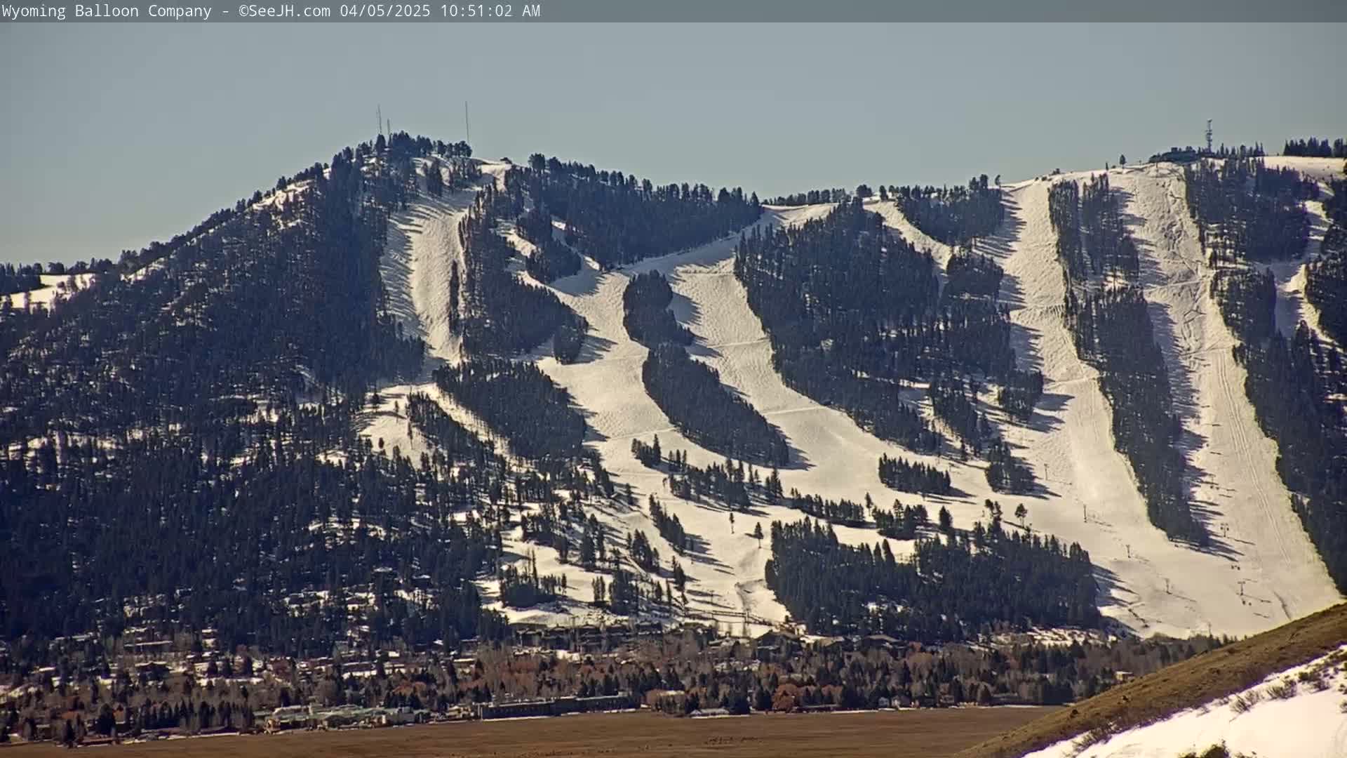 A snow-covered mountain with ski runs and evergreen trees is visible under a clear, sunny sky, with a town situated at its base.
