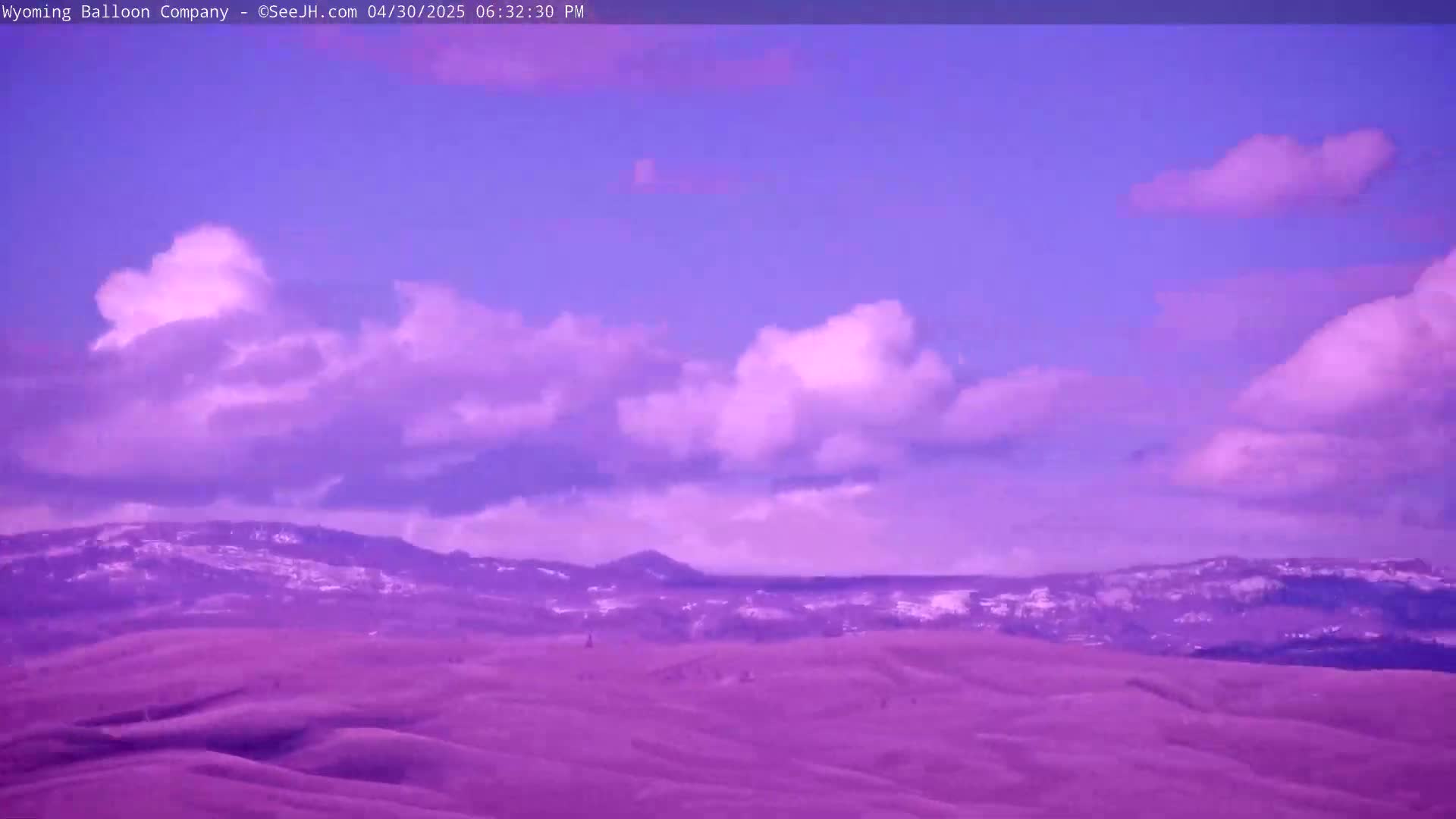Under a mostly clear, purple-hued sky with scattered fluffy clouds, snow-dusted mountains rise behind rolling purple hills.