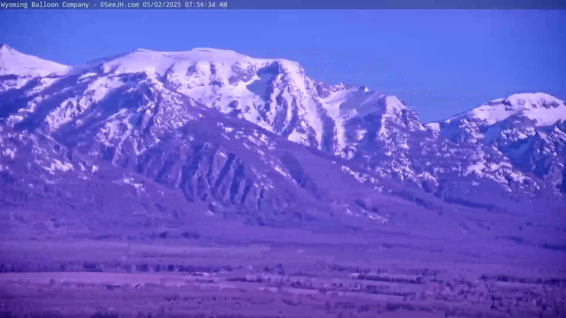 A snow-capped mountain range is visible under a clear blue sky, with a flat, sparsely vegetated valley in the foreground.