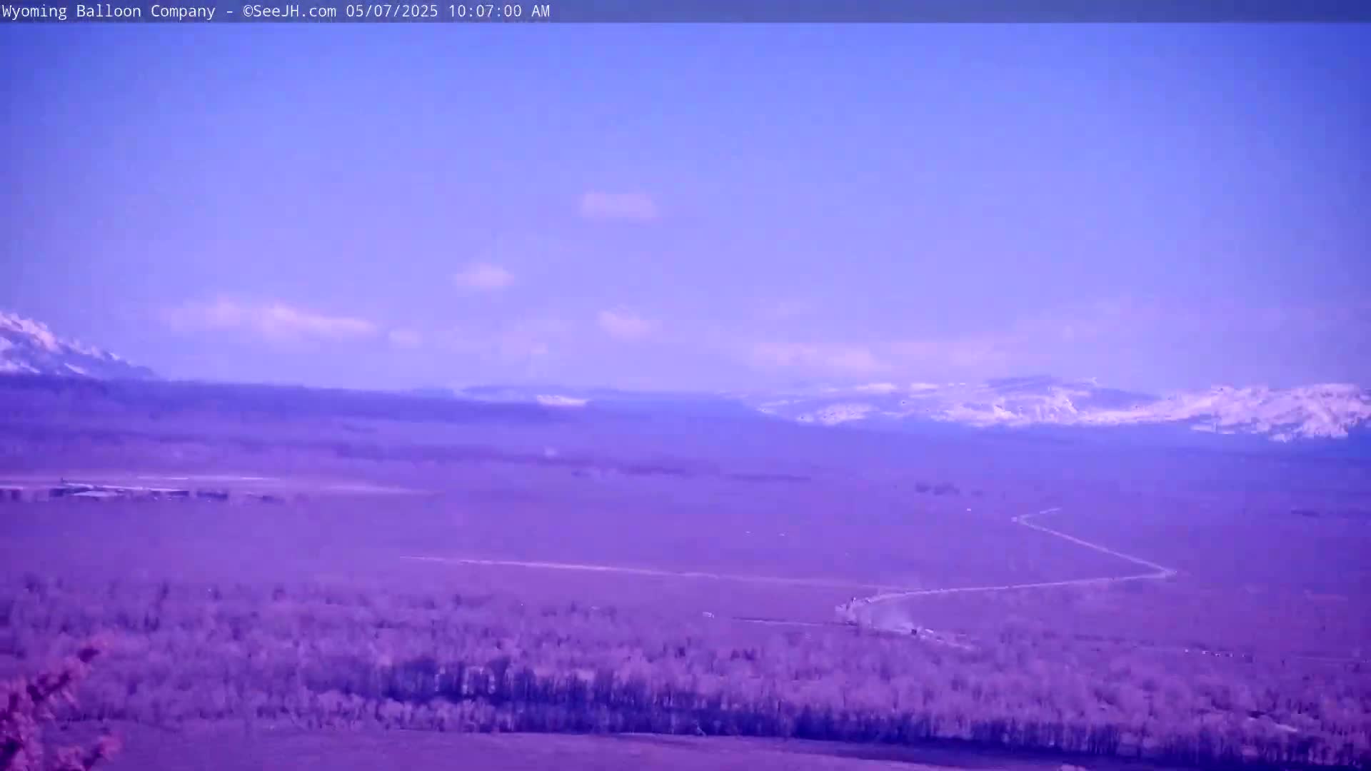 A long, winding road stretches across a flat, purple-tinted landscape toward snow-capped mountains under a mostly clear, light blue sky.