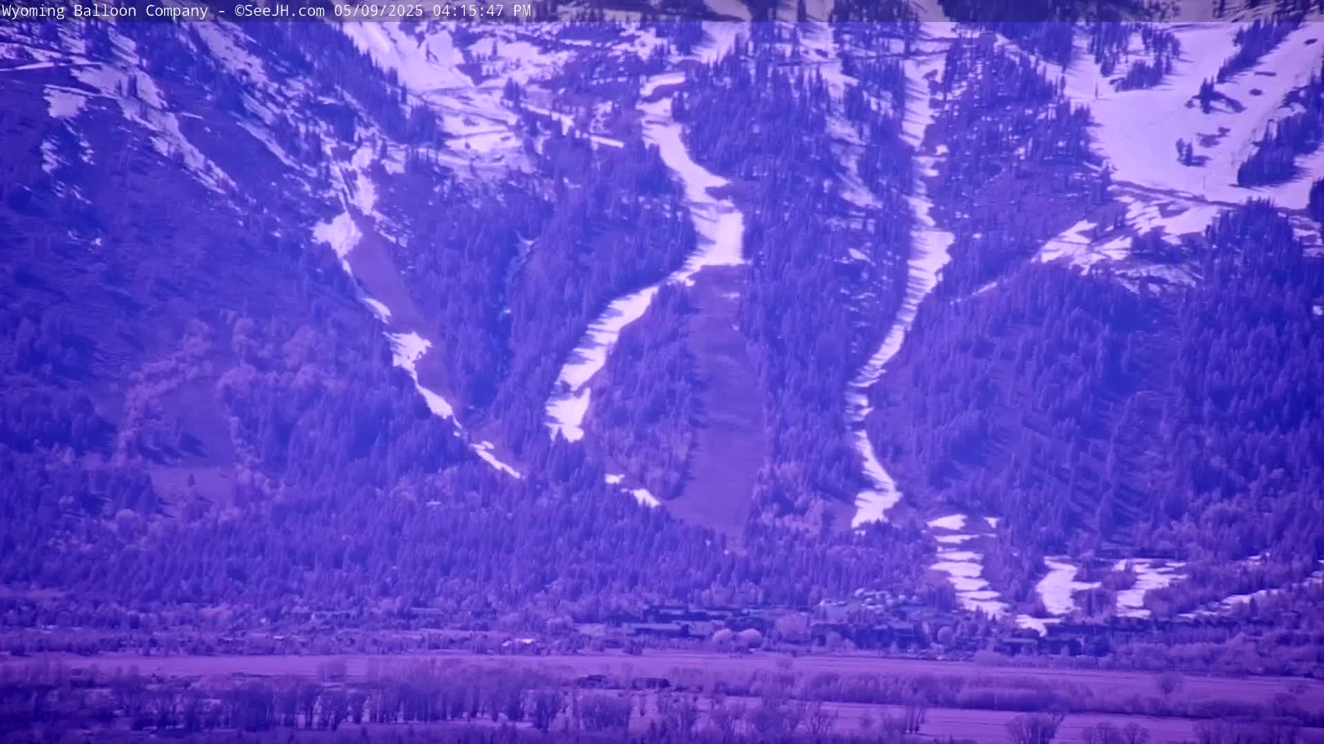 A snow-dusted mountainside with a sparsely populated valley below, viewed on a seemingly clear day.