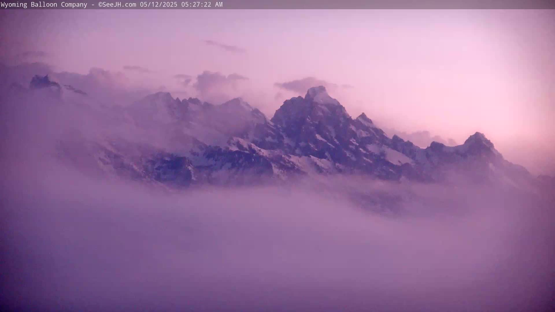 Snow-capped mountains are partially obscured by a thick layer of purple-hued fog under a pale pink sky.