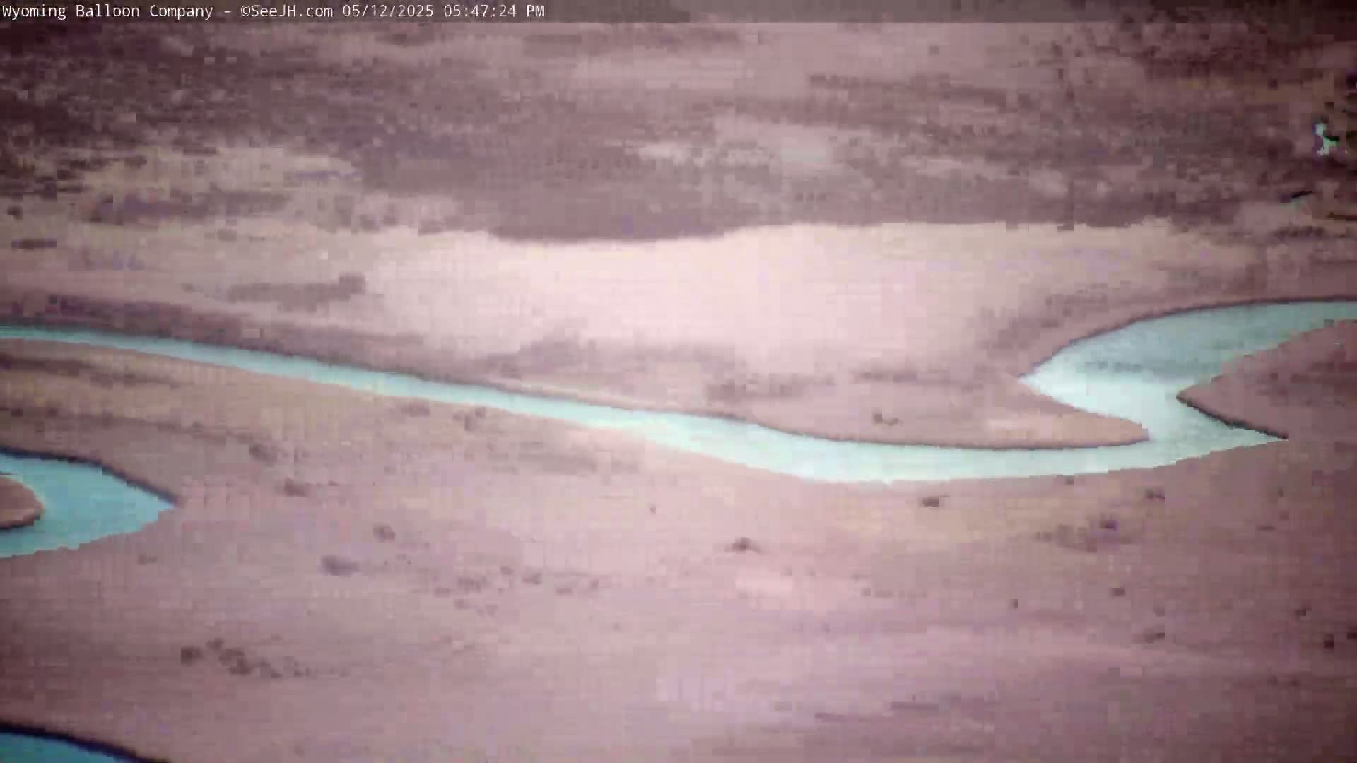 An aerial view shows a light-blue river winding through a pale-brown, sparsely vegetated landscape under a hazy sky.