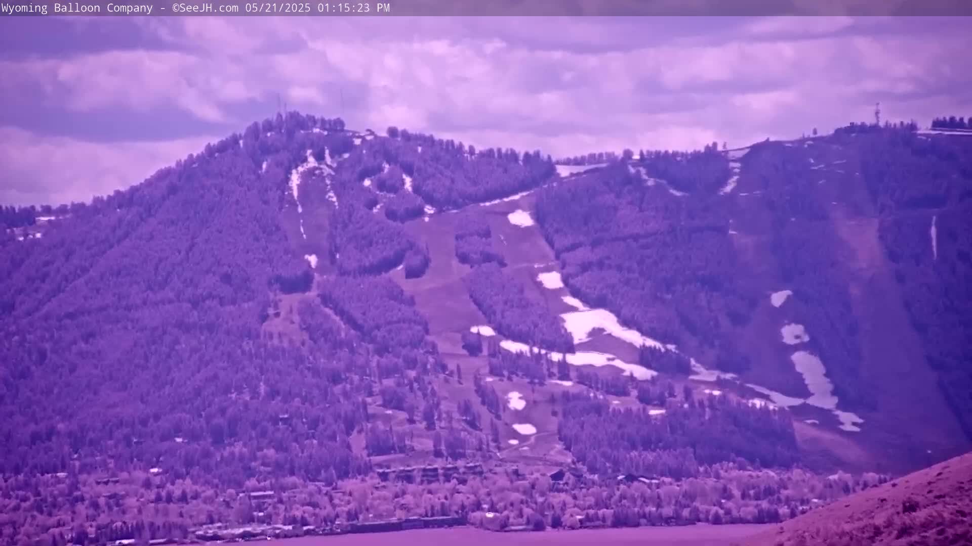 A partly cloudy day reveals a purple-toned mountainside with ski runs and a small town at its base.