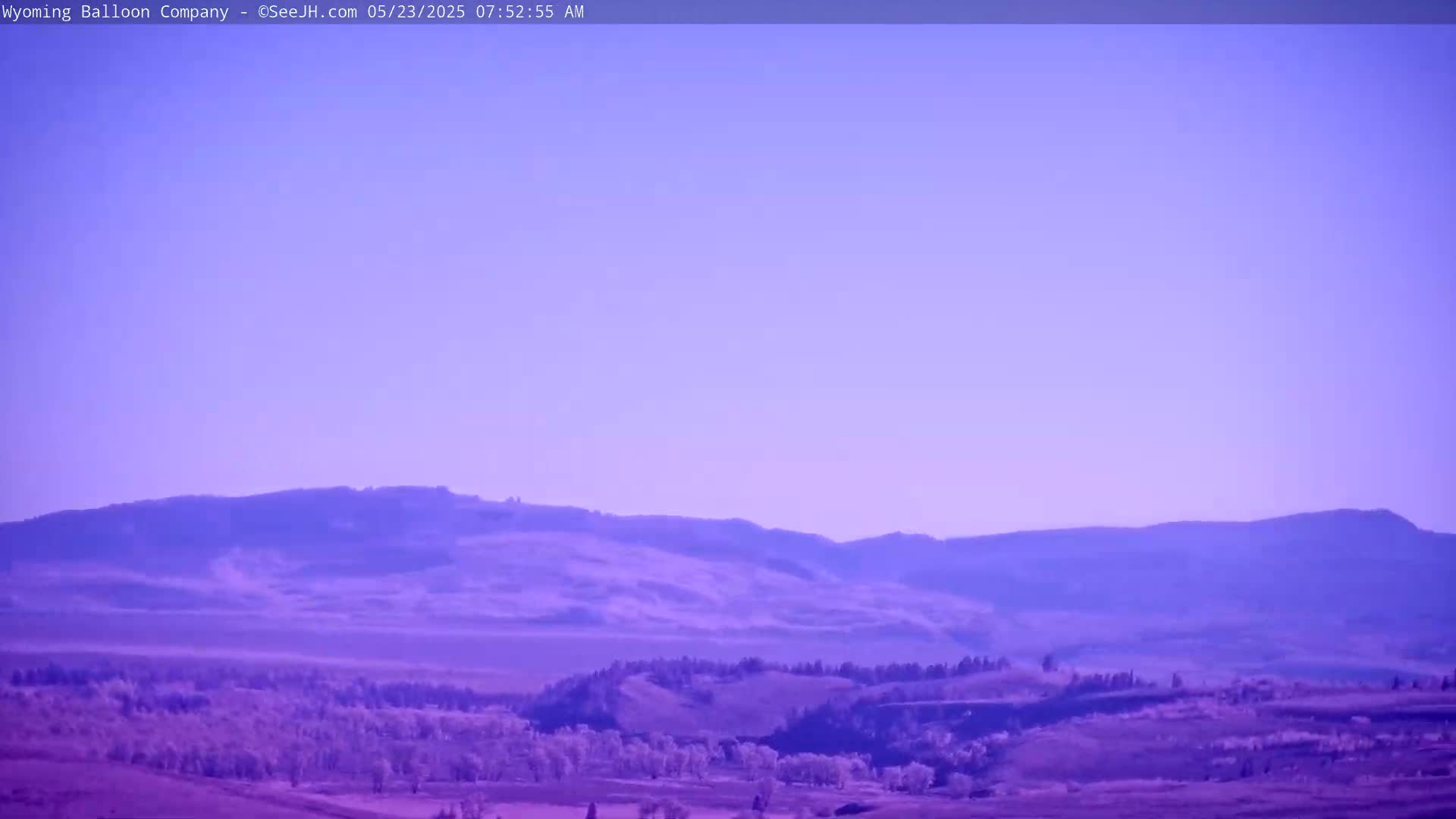 Under a clear, pale purple sky, rolling hills and a flat valley dotted with trees stretch to a distant mountain range.