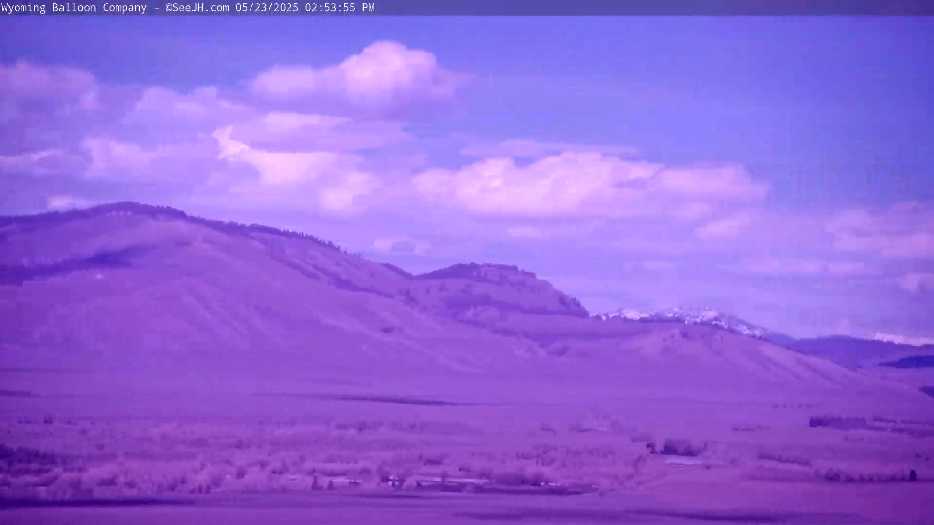 A partly cloudy sky overlooks a purple-toned landscape of rolling hills and distant mountains, some of which have snow-capped peaks.