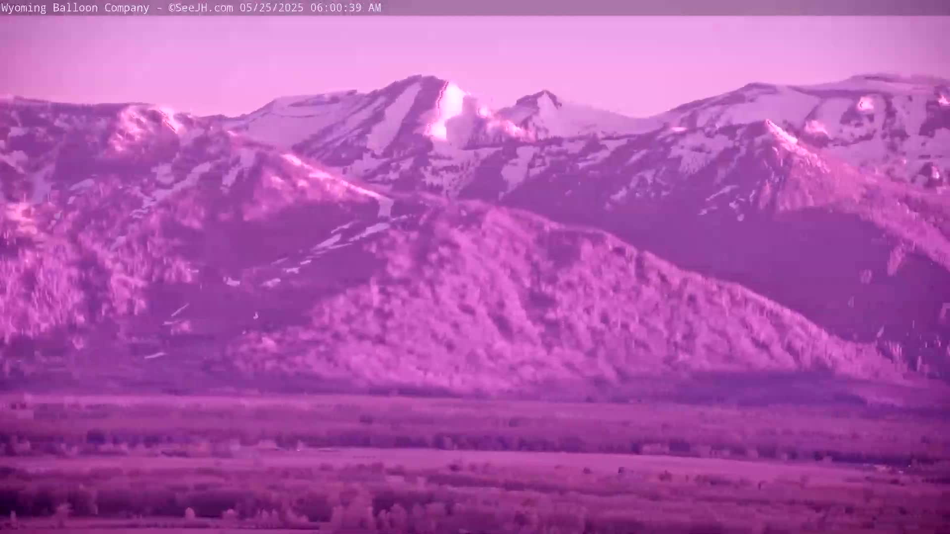 Snow-capped mountains at sunrise are viewed from a distance, over a flat valley.
