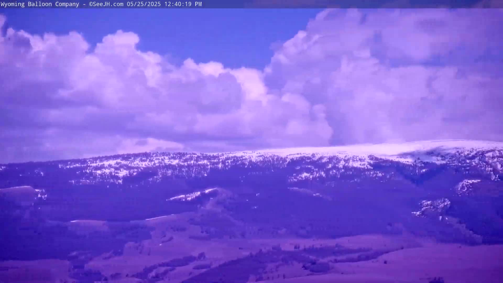 A partly cloudy sky with fluffy white clouds hangs above a snow-dusted mountain range and rolling hills.