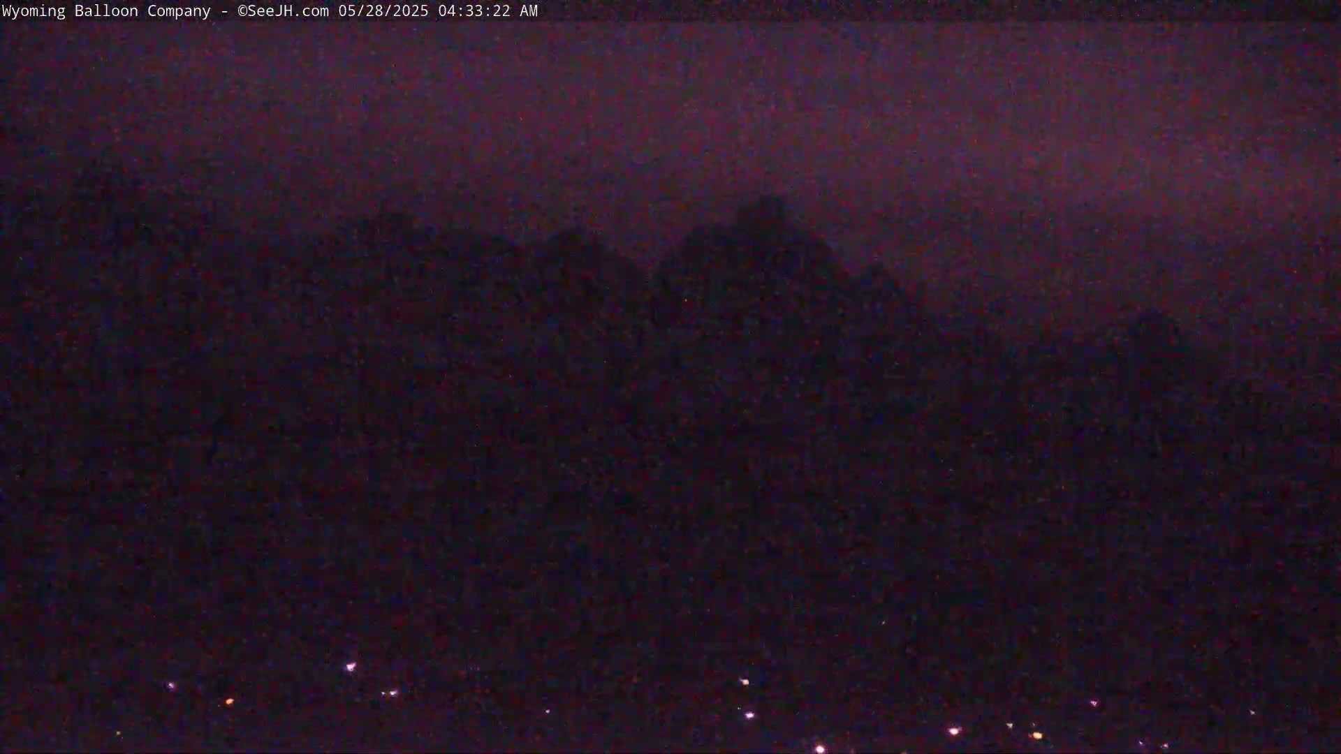 A dark, pre-dawn sky shows silhouetted mountains in the distance, with a few faint lights visible in the foreground.