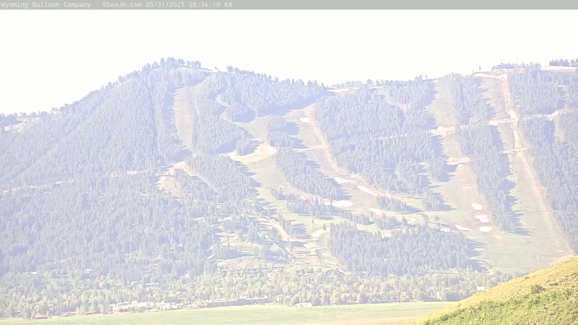 A hazy, long-range view shows a mountainside with numerous ski runs interspersed with evergreen trees, a town nestled at its base, and a flat expanse of land beyond.