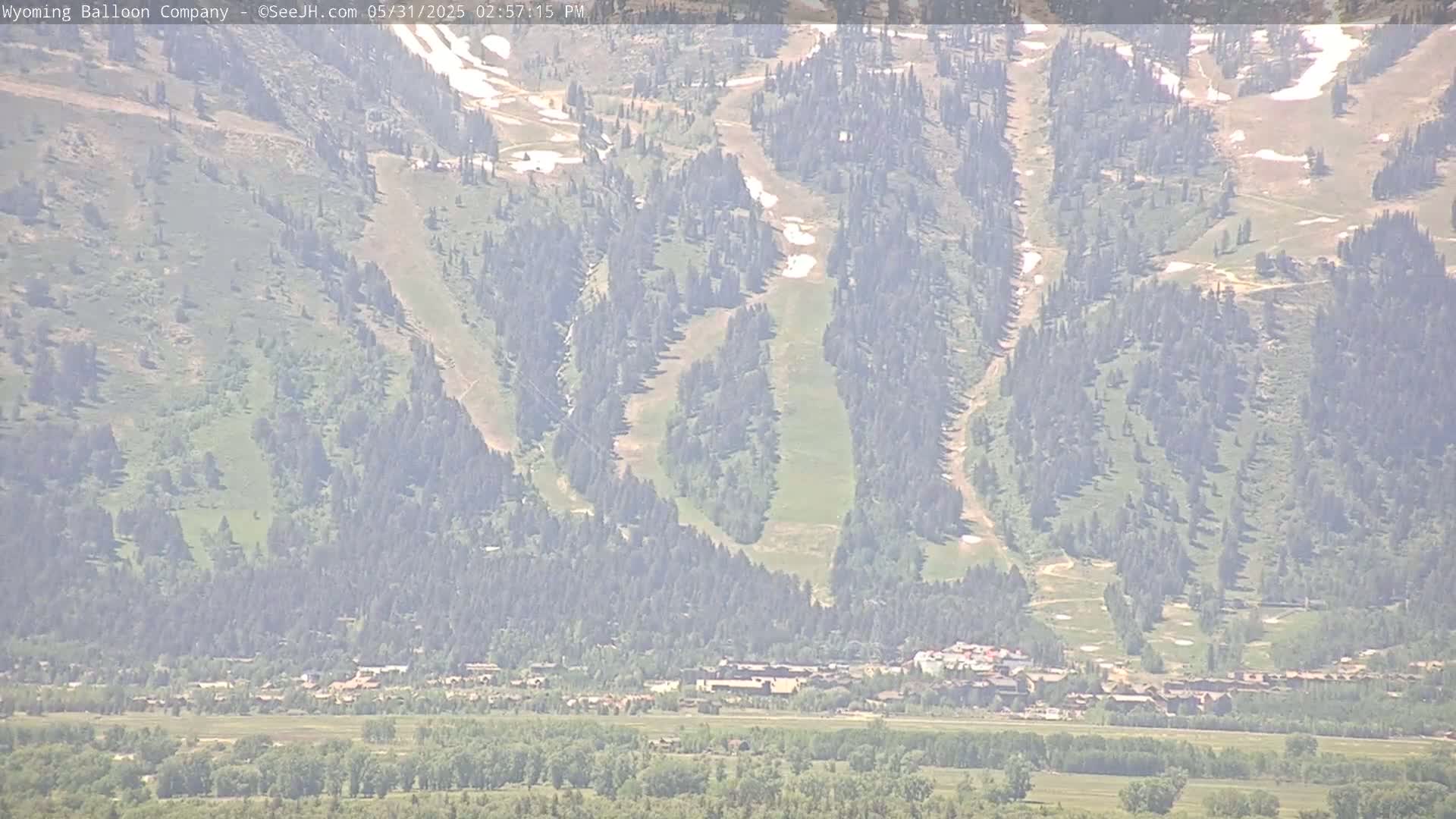 A hazy view shows a mountainside with sparse trees and patches of snow, overlooking a valley with a town and flatlands.