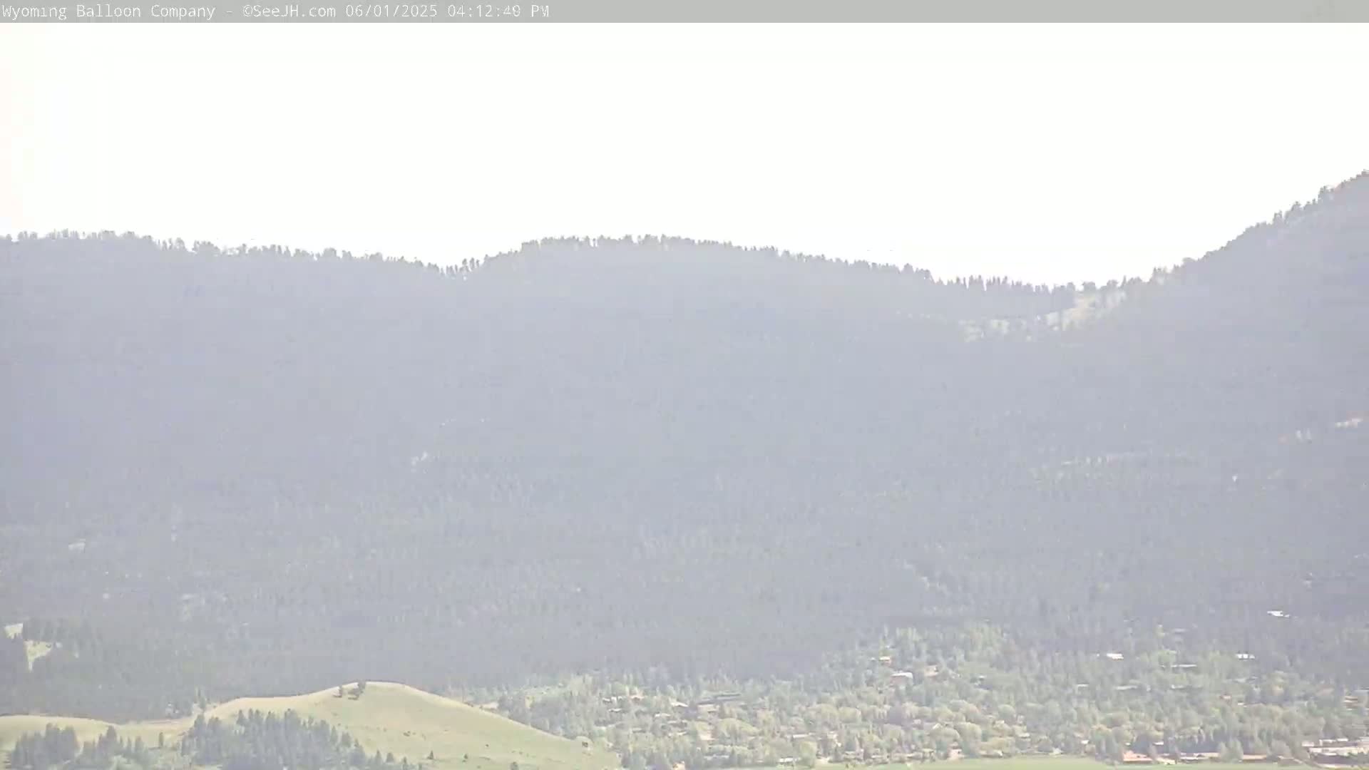 A hazy, sunny view shows a distant town nestled at the base of a large, forested mountain range.