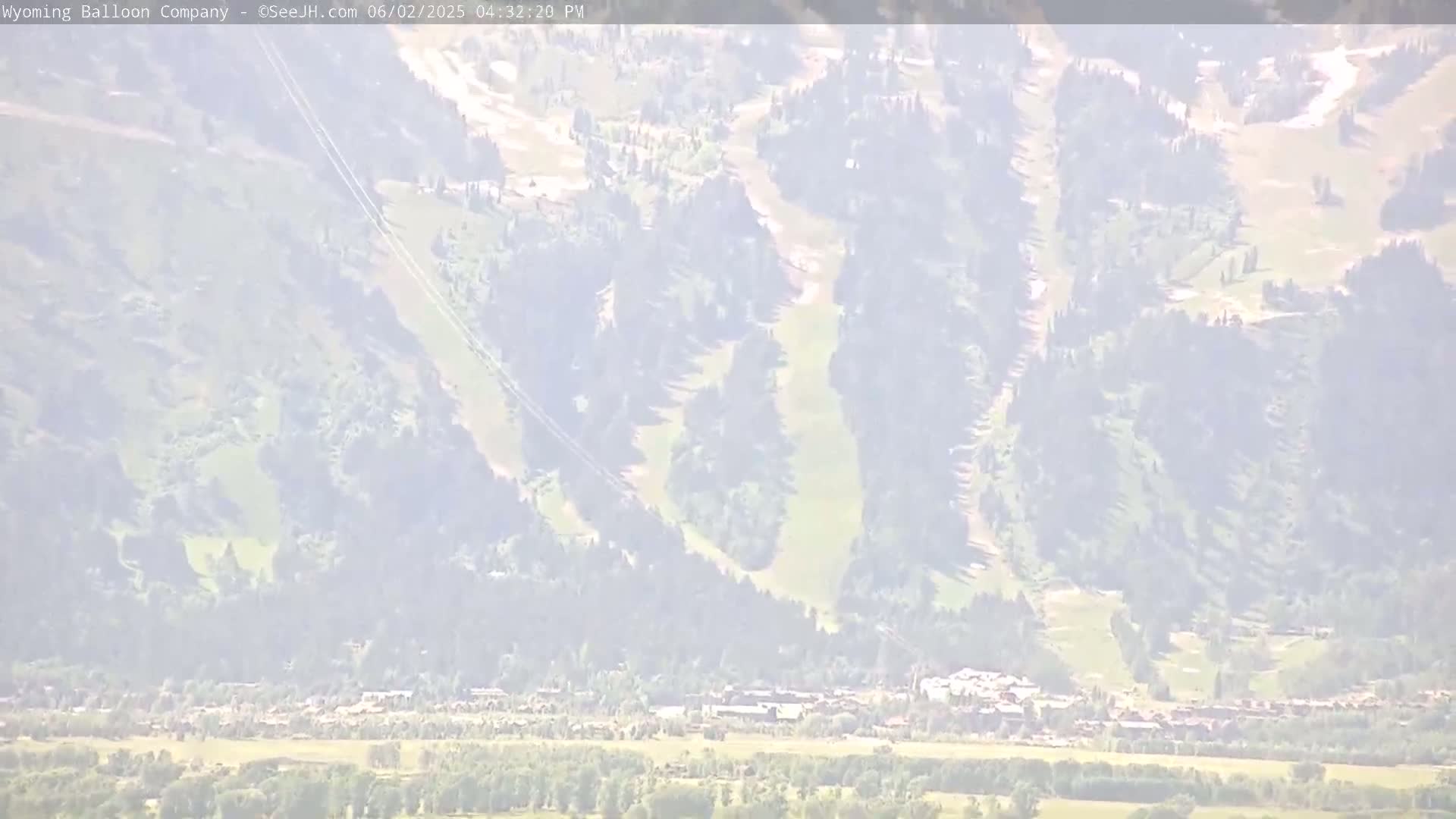 A hazy view shows a town nestled at the base of a mountain with visible ski runs, possibly under a partly sunny sky.