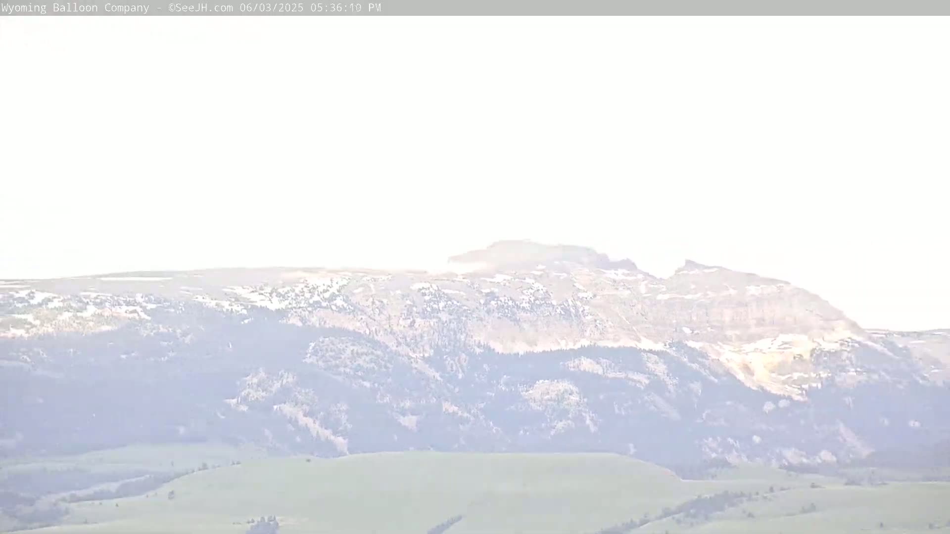 A hazy, distant view shows a mountain range with patches of snow on its peaks and green valleys below.