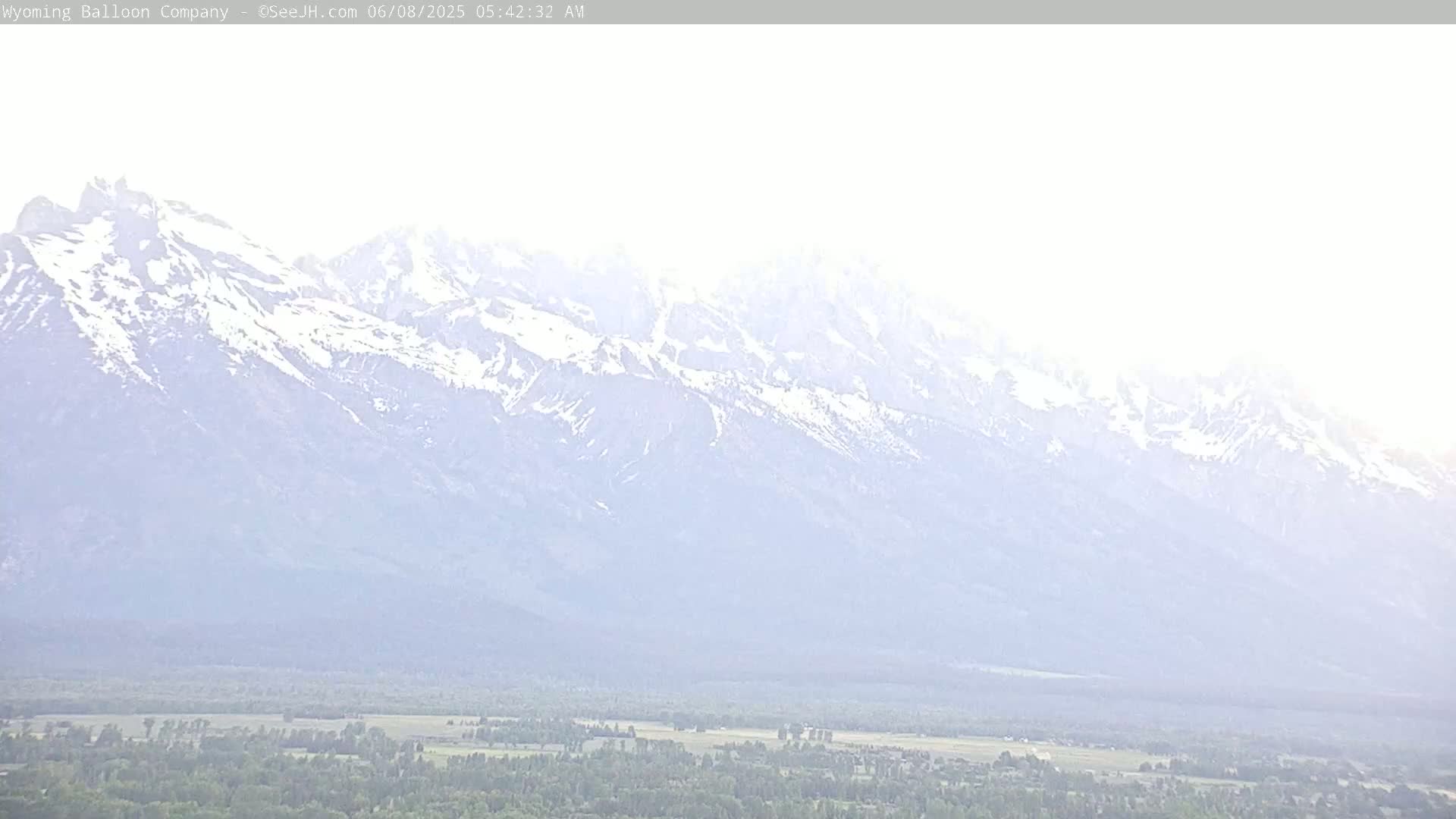 A hazy, bright view shows a snow-capped mountain range overlooking a flat expanse of green fields and trees.