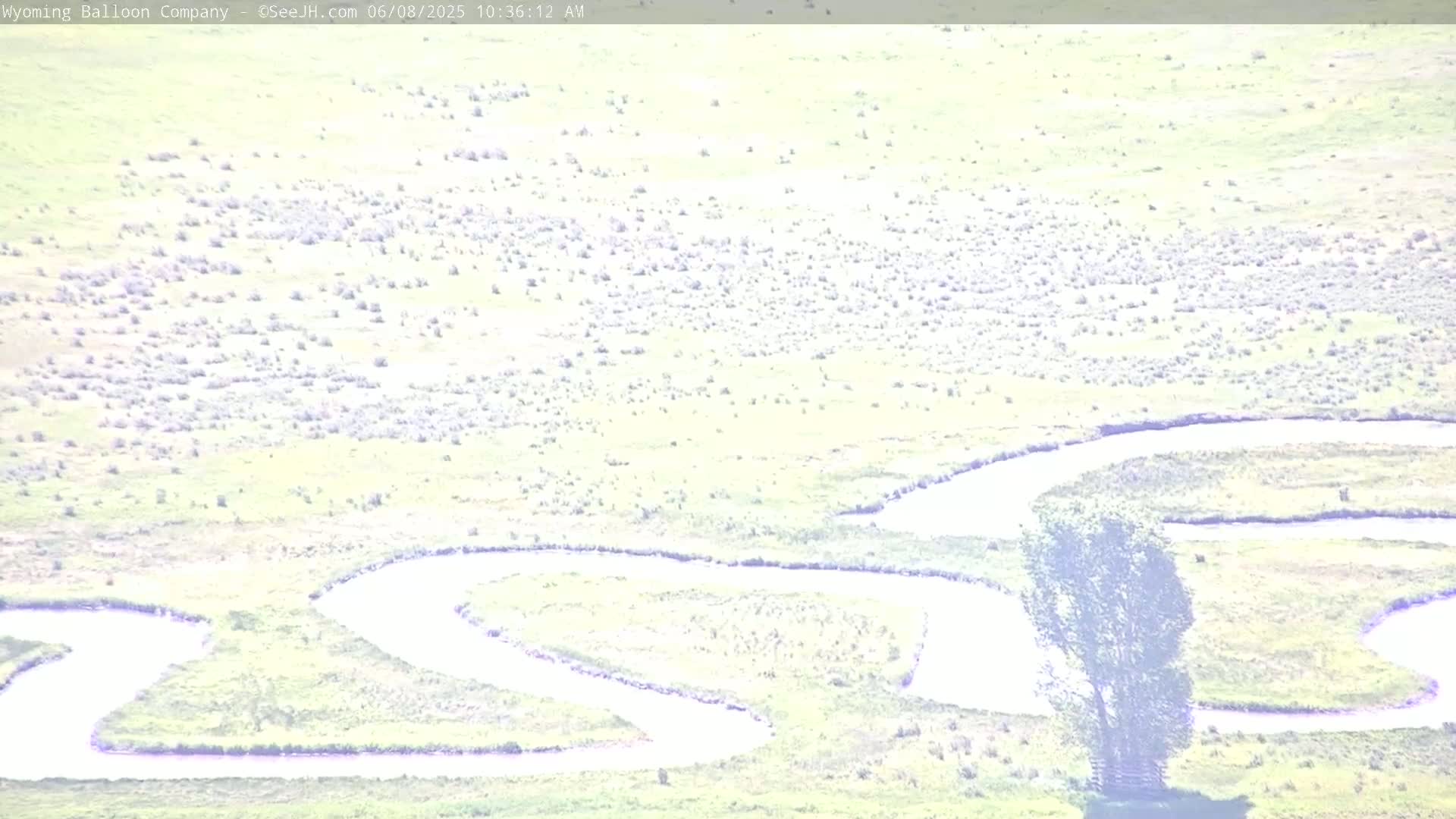 A meandering river flows through a flat, light green field under a bright, sunny sky.