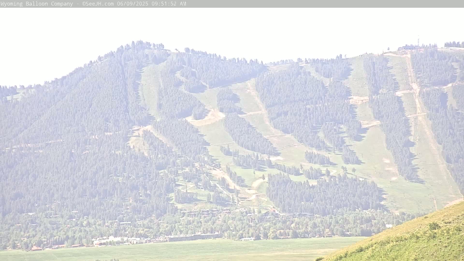 A hazy view shows a forested mountain with groomed ski runs, a small town at its base, and a grassy hillside in the foreground.