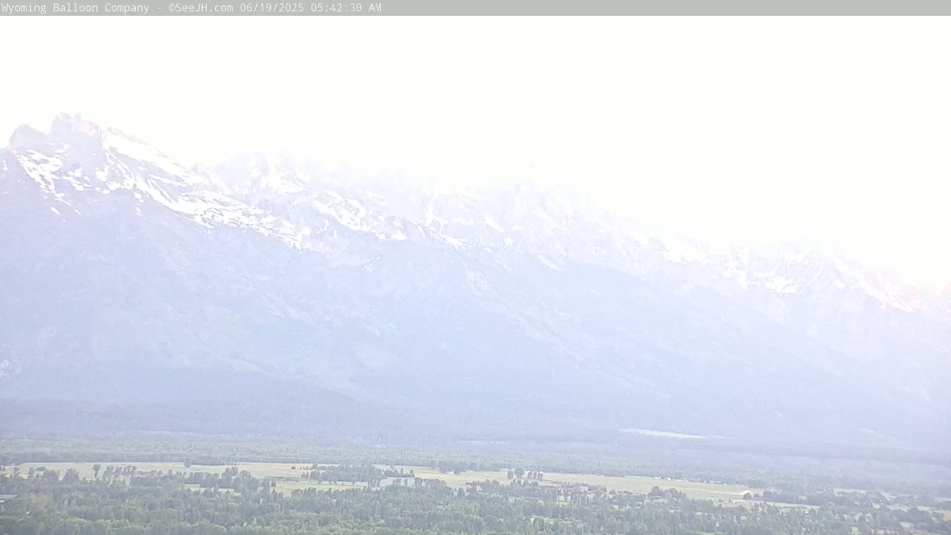 A hazy, light-colored mountain range with snow-capped peaks is visible in the distance above a flat, green valley.