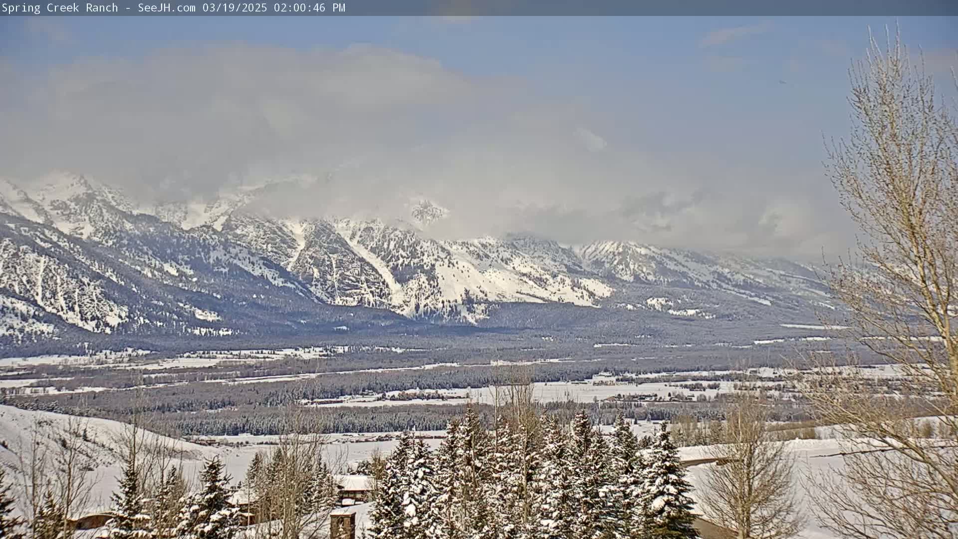 A snow-covered valley with a snow-capped mountain range in the background under a partly cloudy sky.
