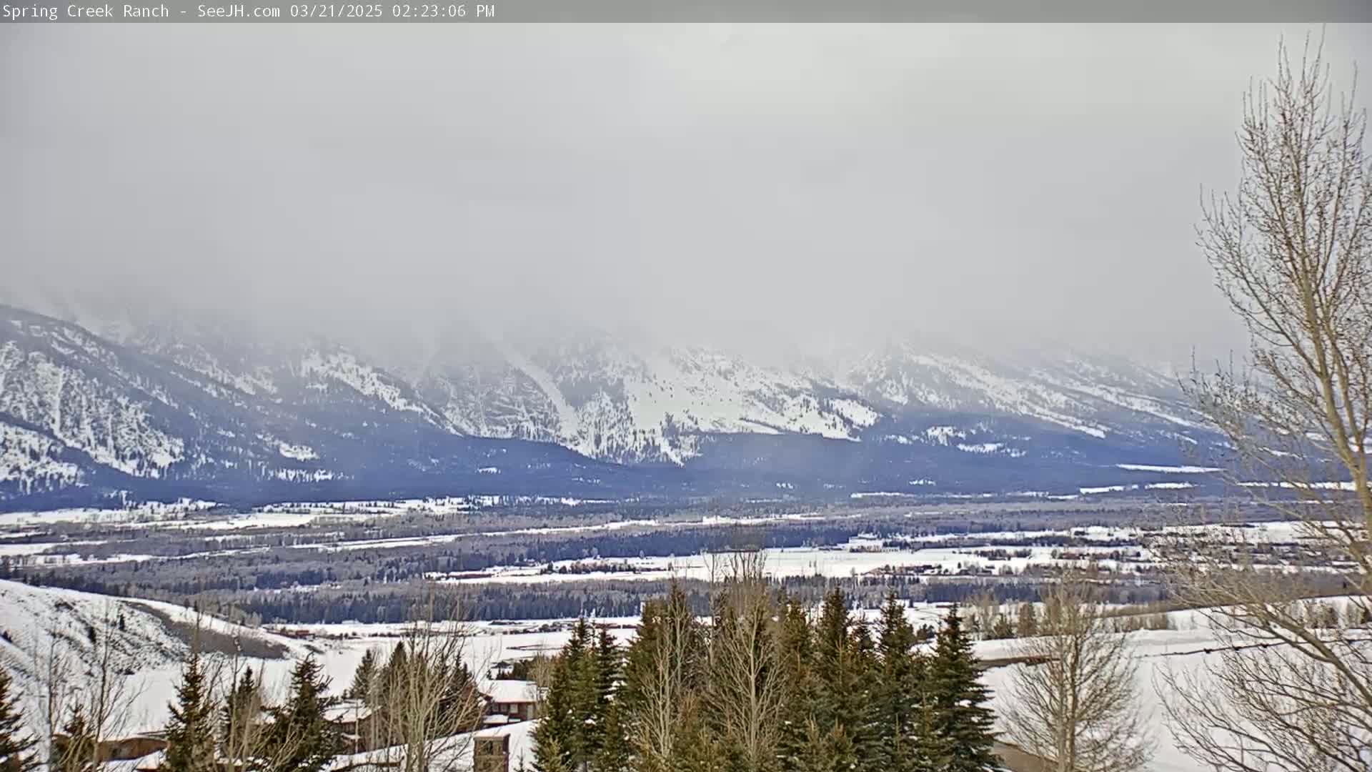 A hazy, snowy landscape reveals snow-capped mountains in the distance, with a valley of snow-covered trees and buildings in the foreground.