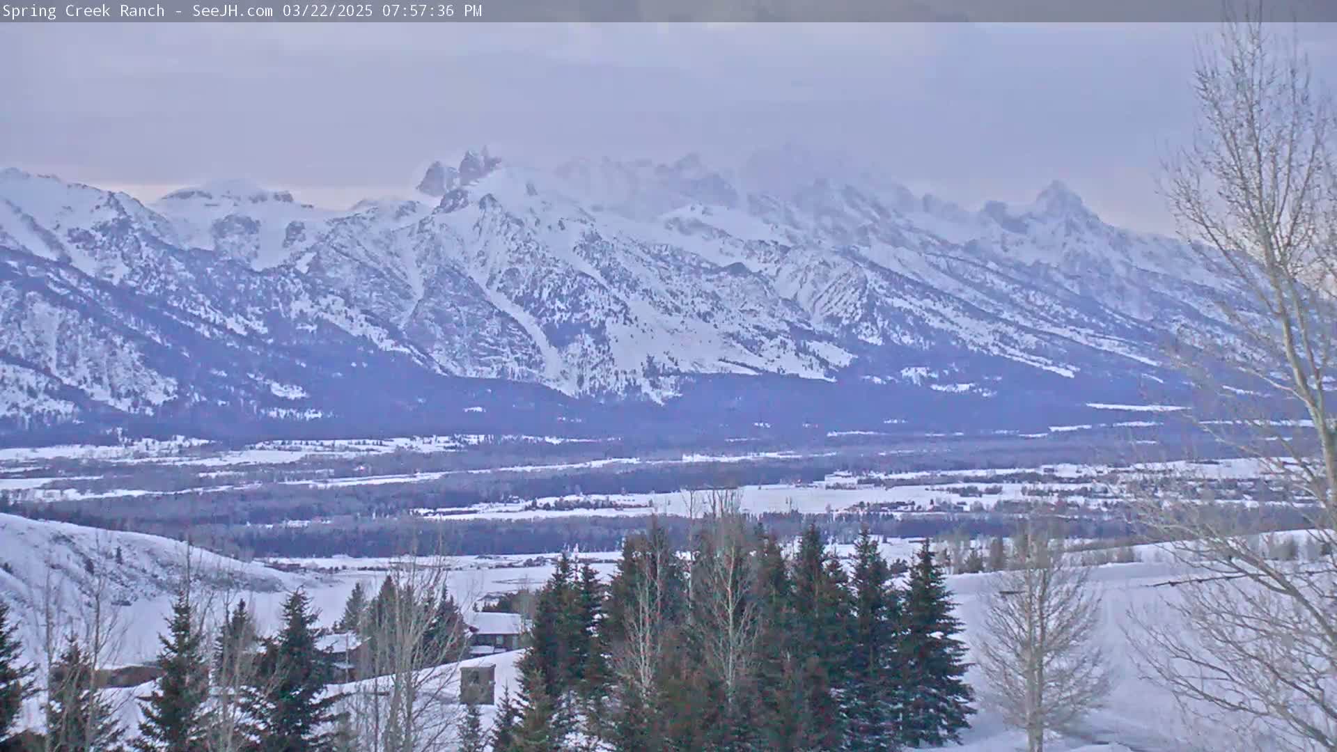 A snow-covered valley stretches out before a range of snow-capped mountains under a hazy sky.