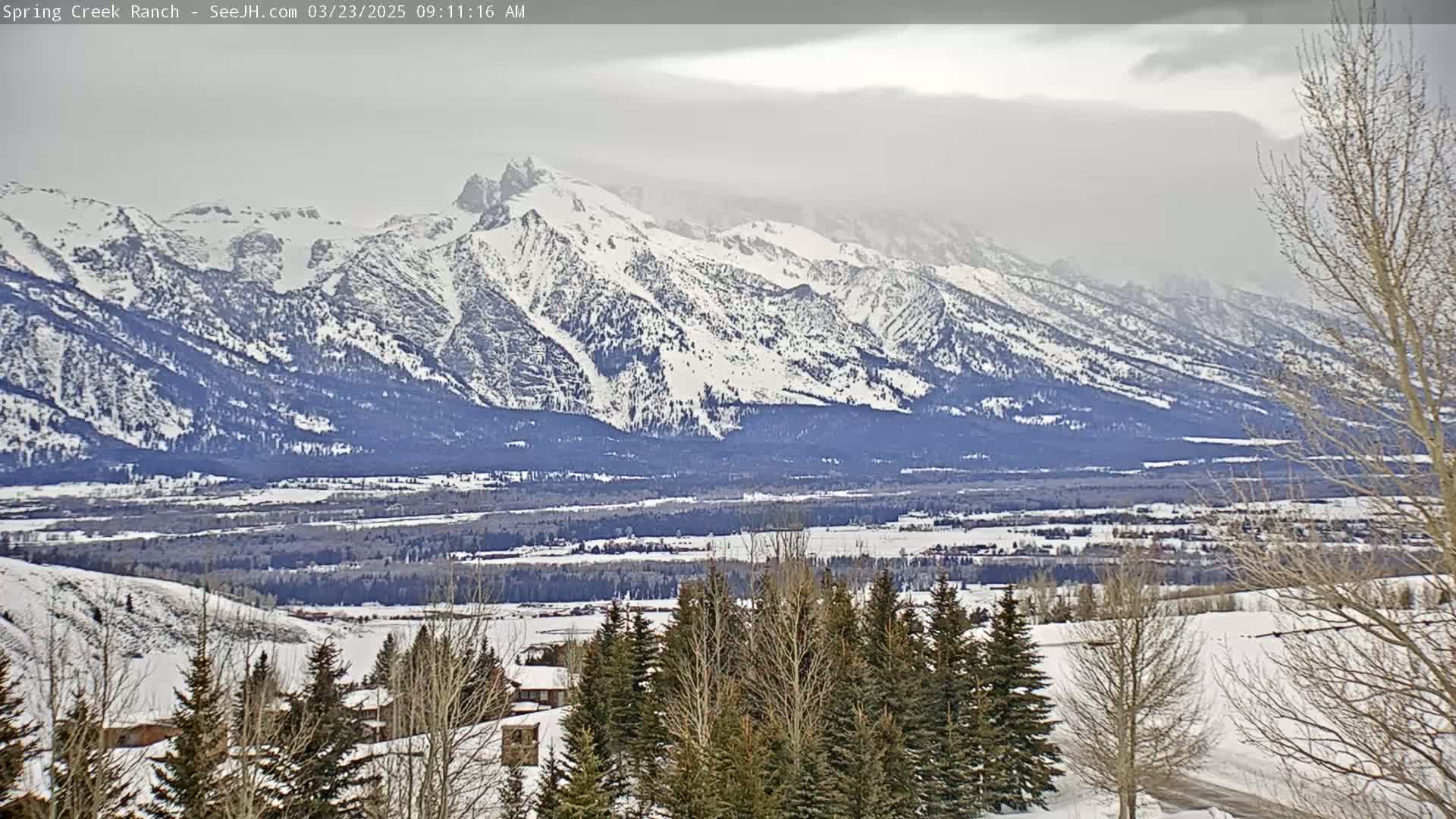 A snow-covered valley stretches out before a large, snow-capped mountain range under a cloudy sky.