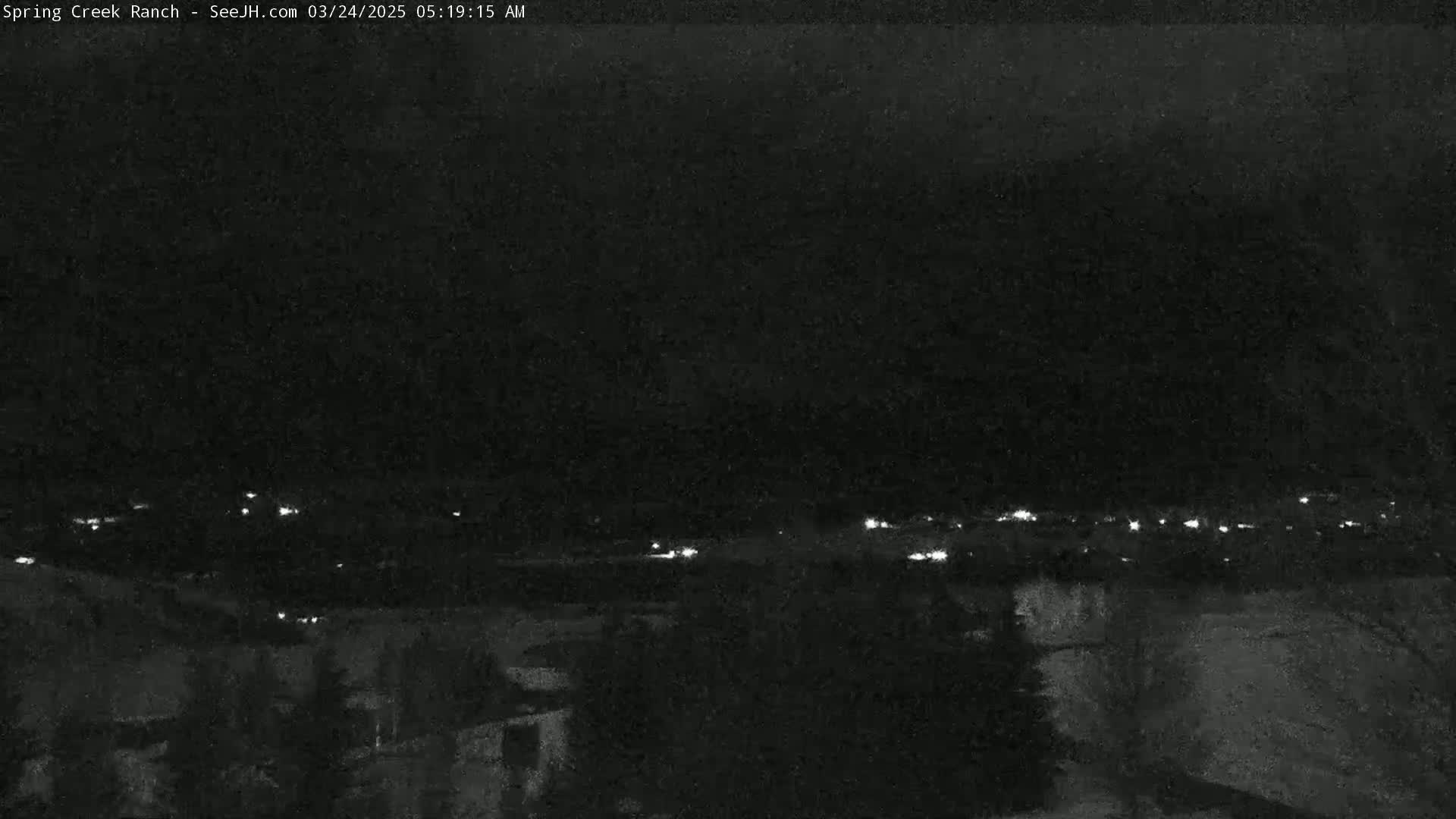 A nighttime, low-light view shows a distant line of lights possibly from a town or city, with dark, shadowy terrain in the foreground.