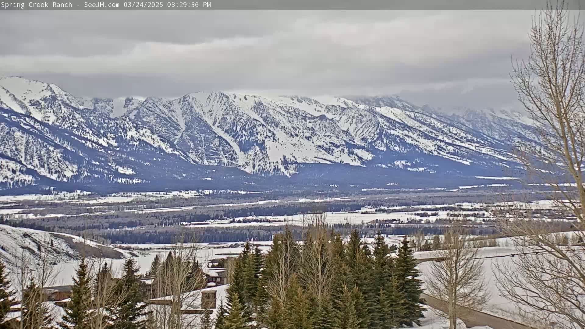 A snow-covered valley, dotted with evergreens and buildings, stretches out before a range of snow-capped mountains under a cloudy sky.