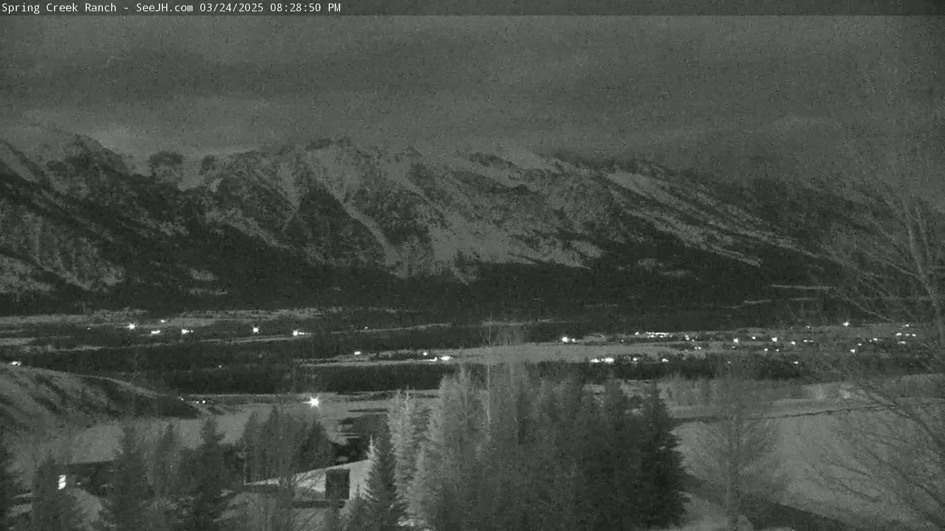A nighttime view shows snow-covered mountains in the distance, a valley with scattered lights below, and dark trees in the foreground under a cloudy sky.